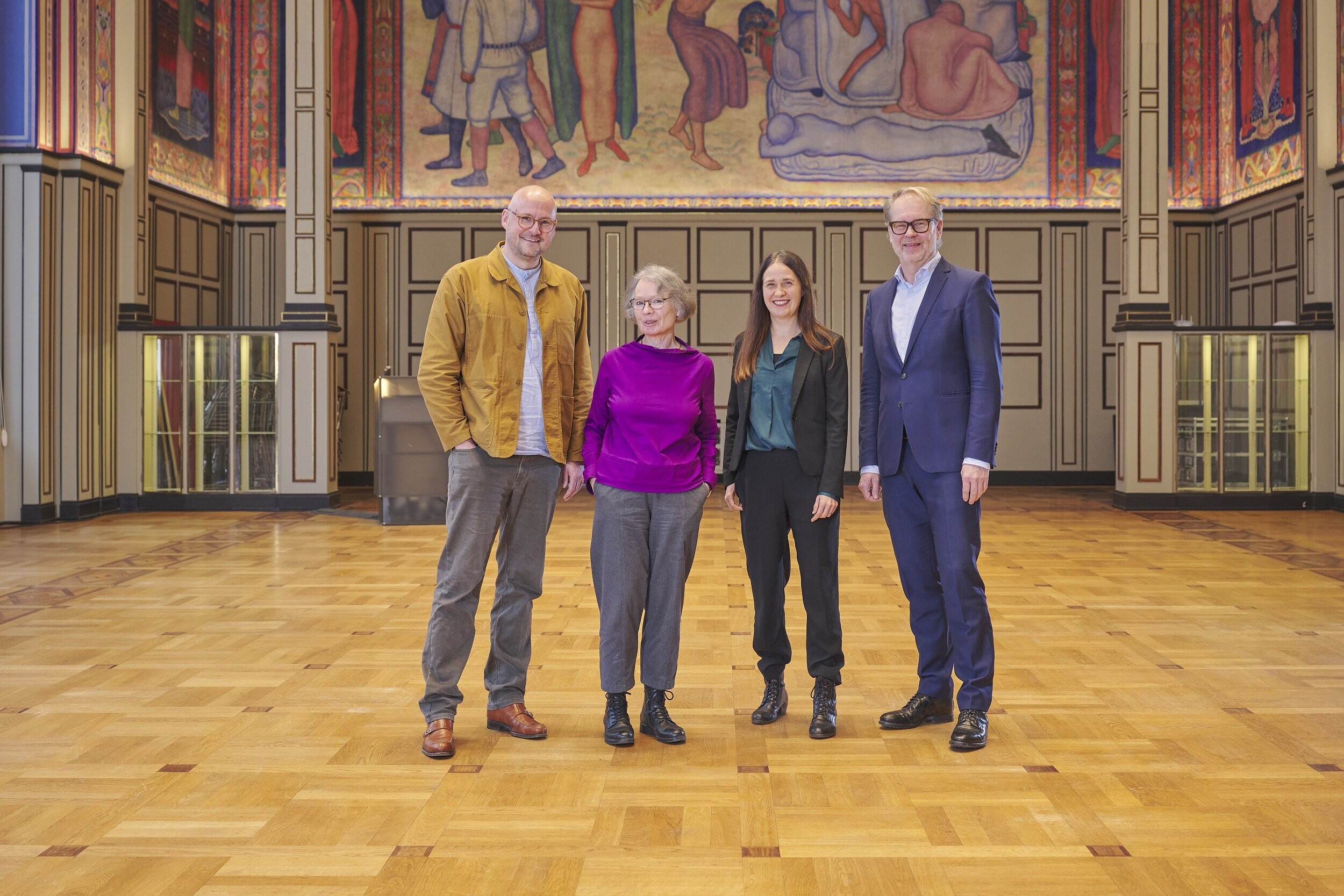 Four people are standing for a group photo in a large, ornate hall with colorful wall murals and a parquet floor. Two men are on the far left and far right, and two women stand in the middle. They are all smiling at the camera and wearing a mix of semi-formal and formal clothing.