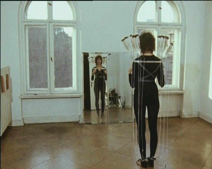 A person in a black, form-fitting outfit stands in a bright room in front of a mirror, wearing a construction made of long threads and brushes; the mirror reflects the scene.