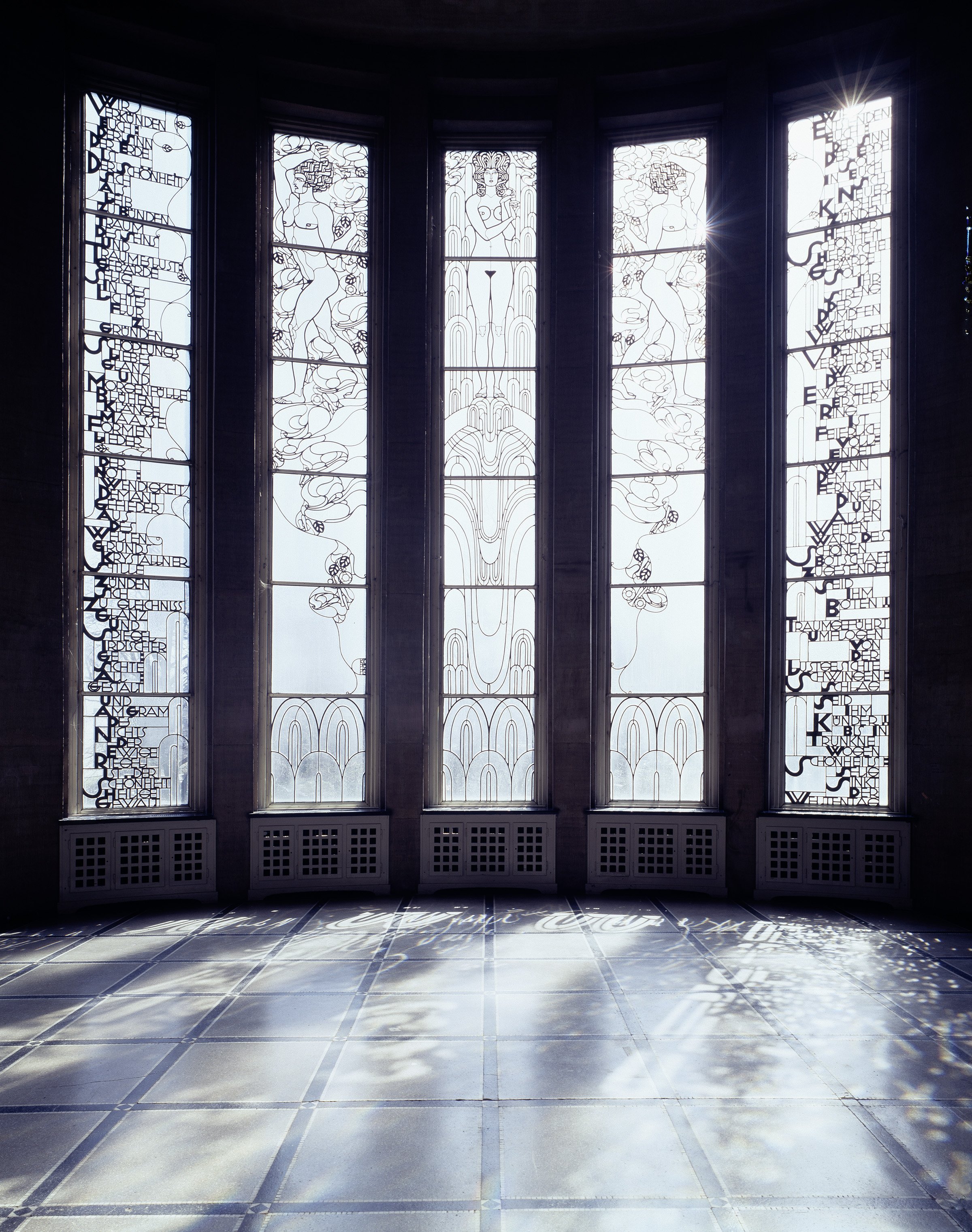 Room featuring five tall, ornate windows with delicate line drawings of figures, plants, and text. Sunlight casts patterned shadows of the window designs onto the floor. The atmosphere is calm and filled with light.