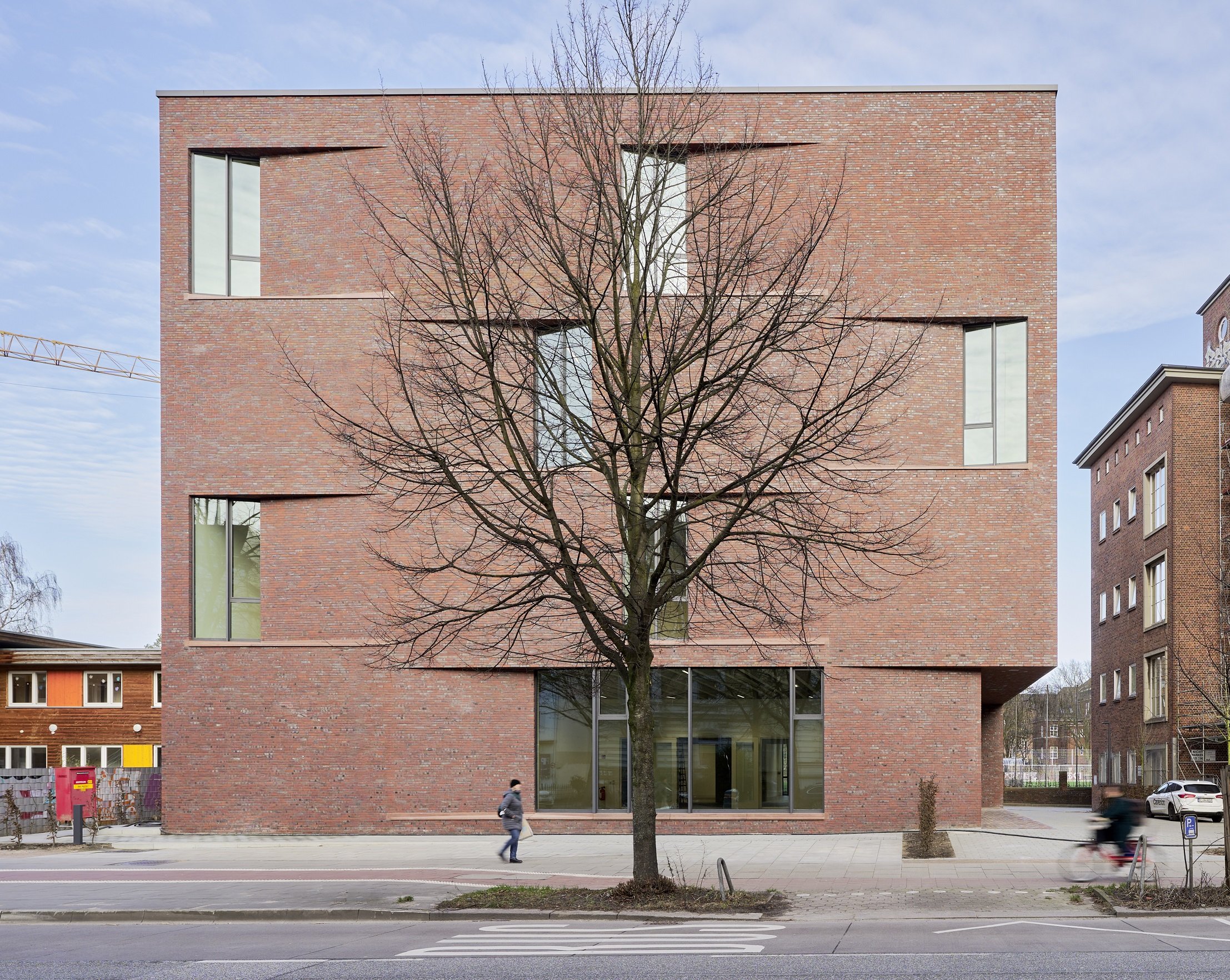 Modern brick façade of a cubic building with narrow, irregularly arranged windows; in the foreground stands a leafless tree, a person is walking on the sidewalk, and a bicycle passes by.