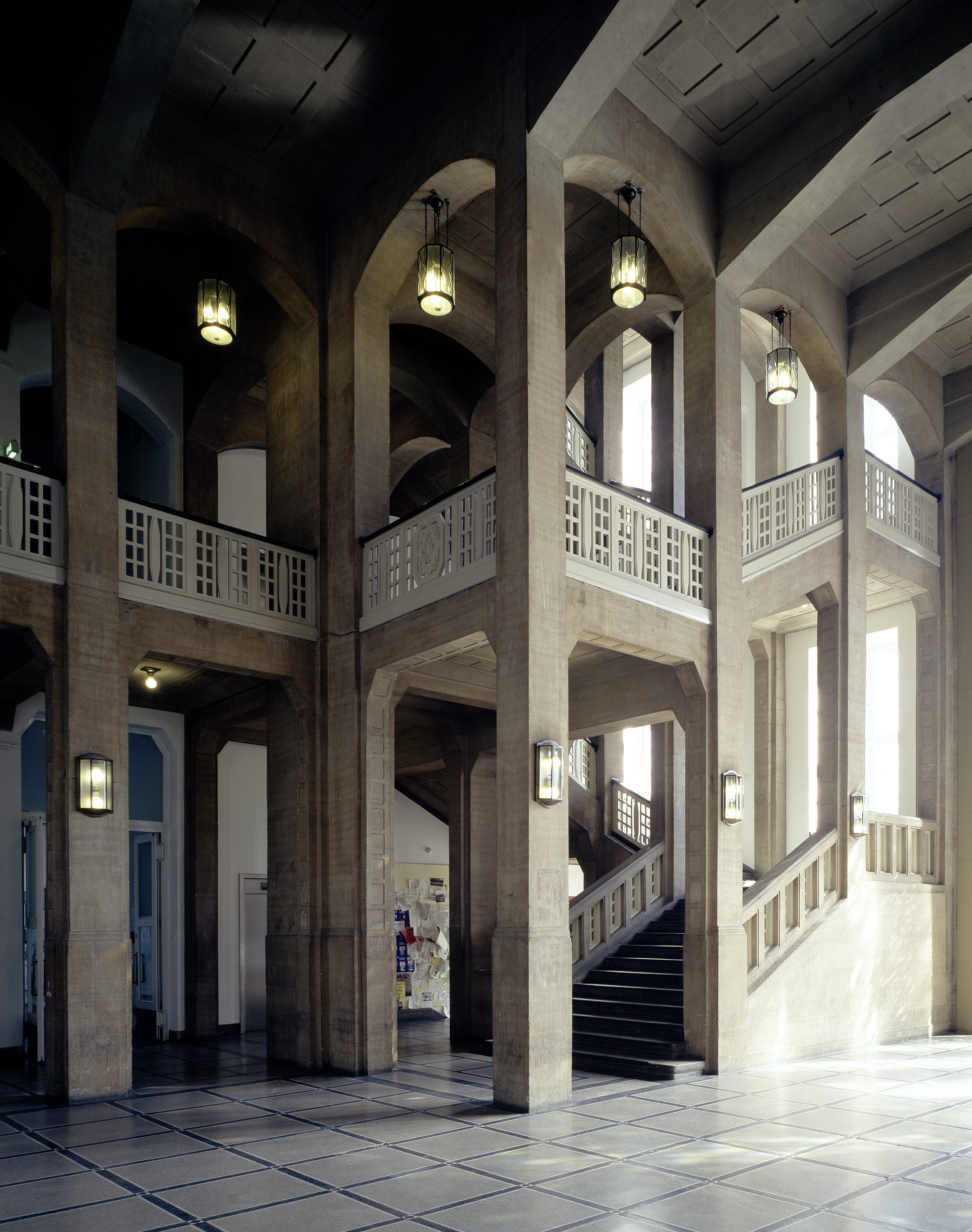 A tall, light-filled staircase hall featuring massive pillars and wide arches. Multiple stairways lead upward, bordered by pale railings with geometric patterns. Hanging lantern lights and natural daylight highlight the architectural structure.