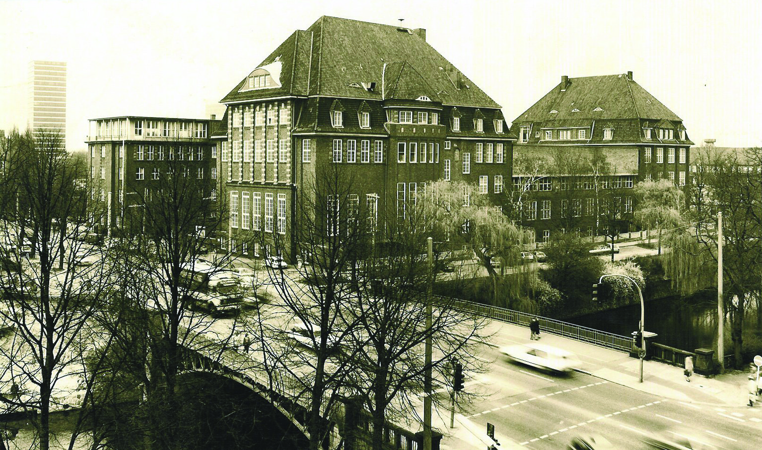 Historic black-and-white photograph of a large brick building complex in an early 20th-century architectural style, located at a busy intersection, with a bridge and leafless trees in the foreground.