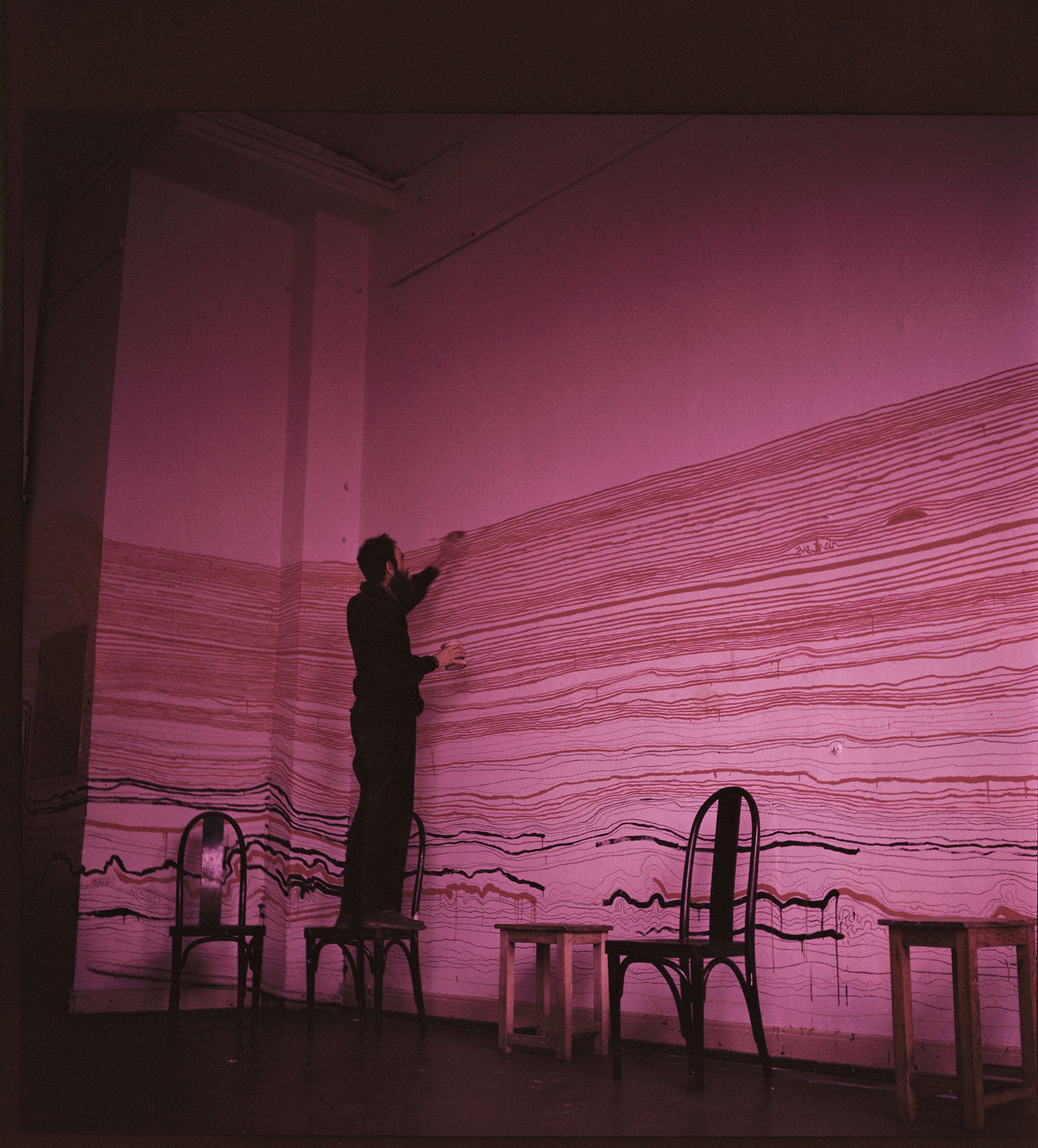 A person stands on a chair, drawing horizontal, wave-like lines on a tall wall illuminated by pink light. Several chairs and stools are placed in front of the wall. The scene appears artistic and experimental.