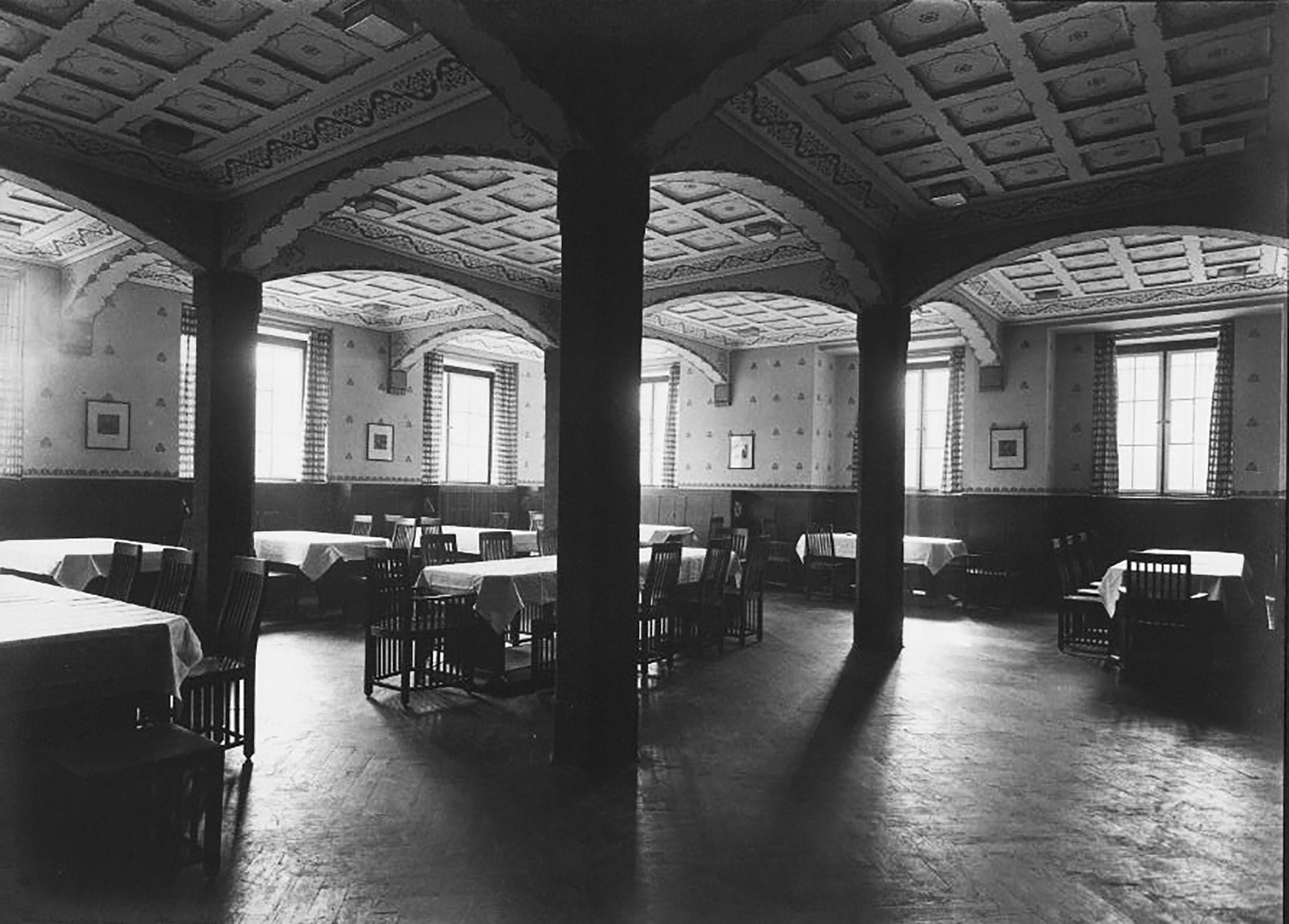 Historic black-and-white photograph of a dining or common room with a vaulted, coffered ceiling supported by columns; several tables with white tablecloths and wooden chairs are arranged throughout the space, illuminated by daylight from tall windows.