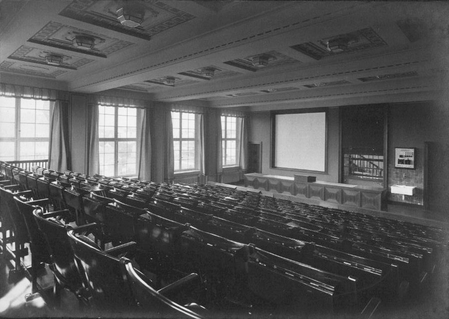 Historic black-and-white photograph of a lecture hall with rows of fixed wooden seats; at the front, a large projection screen and podium, with daylight entering through tall side windows.