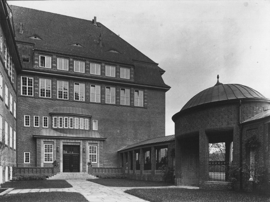 Historic black-and-white photograph of a multi-story brick building with a steep roof and symmetrical windows; to the right stands a round pavilion with a domed roof and open colonnade, connected by a covered walkway.
