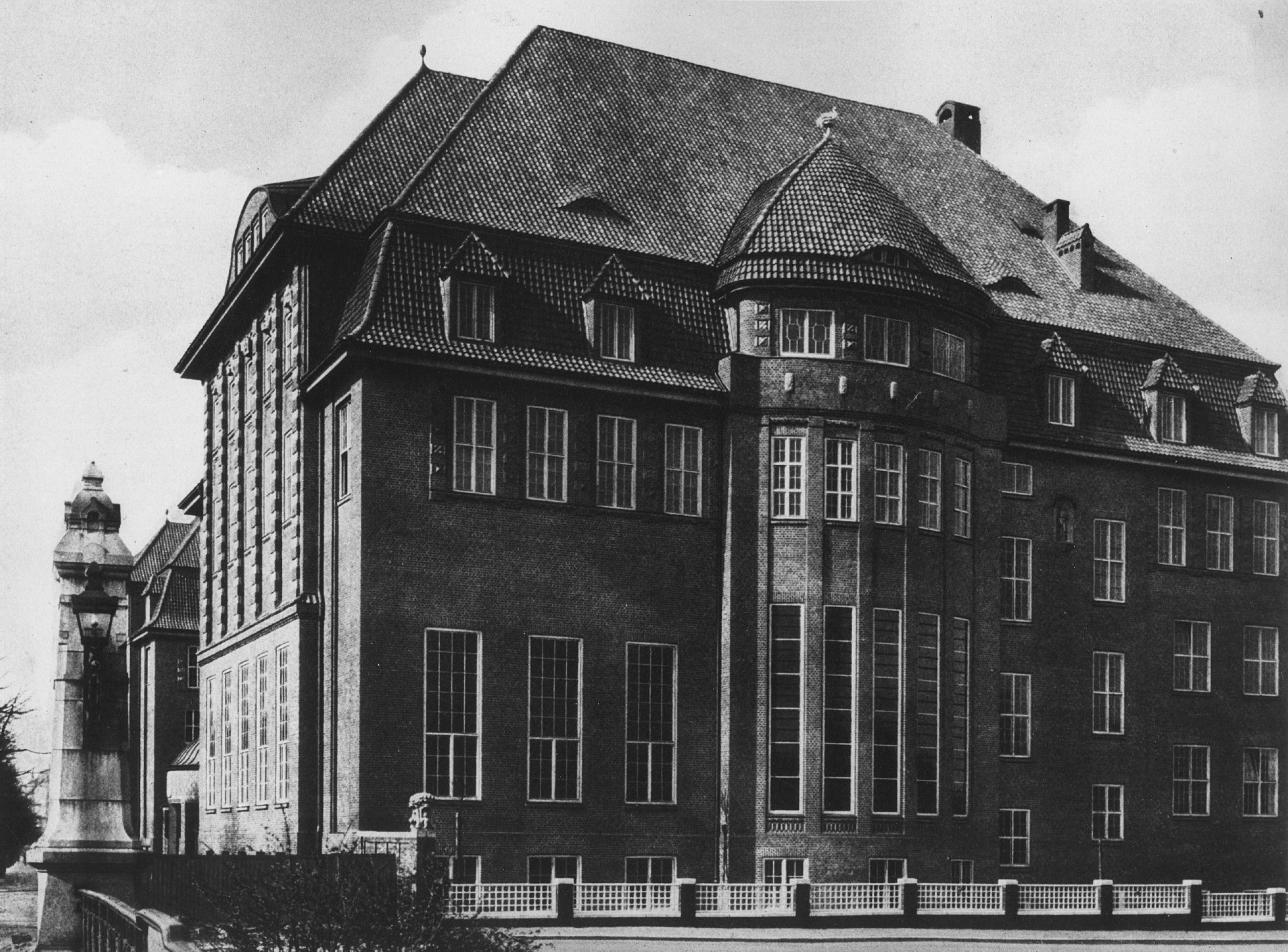 Historic black-and-white photograph of a large brick building with a steep tiled roof, bay windows, and tall multi-pane windows; in the foreground, a bridge railing and a stone lantern.