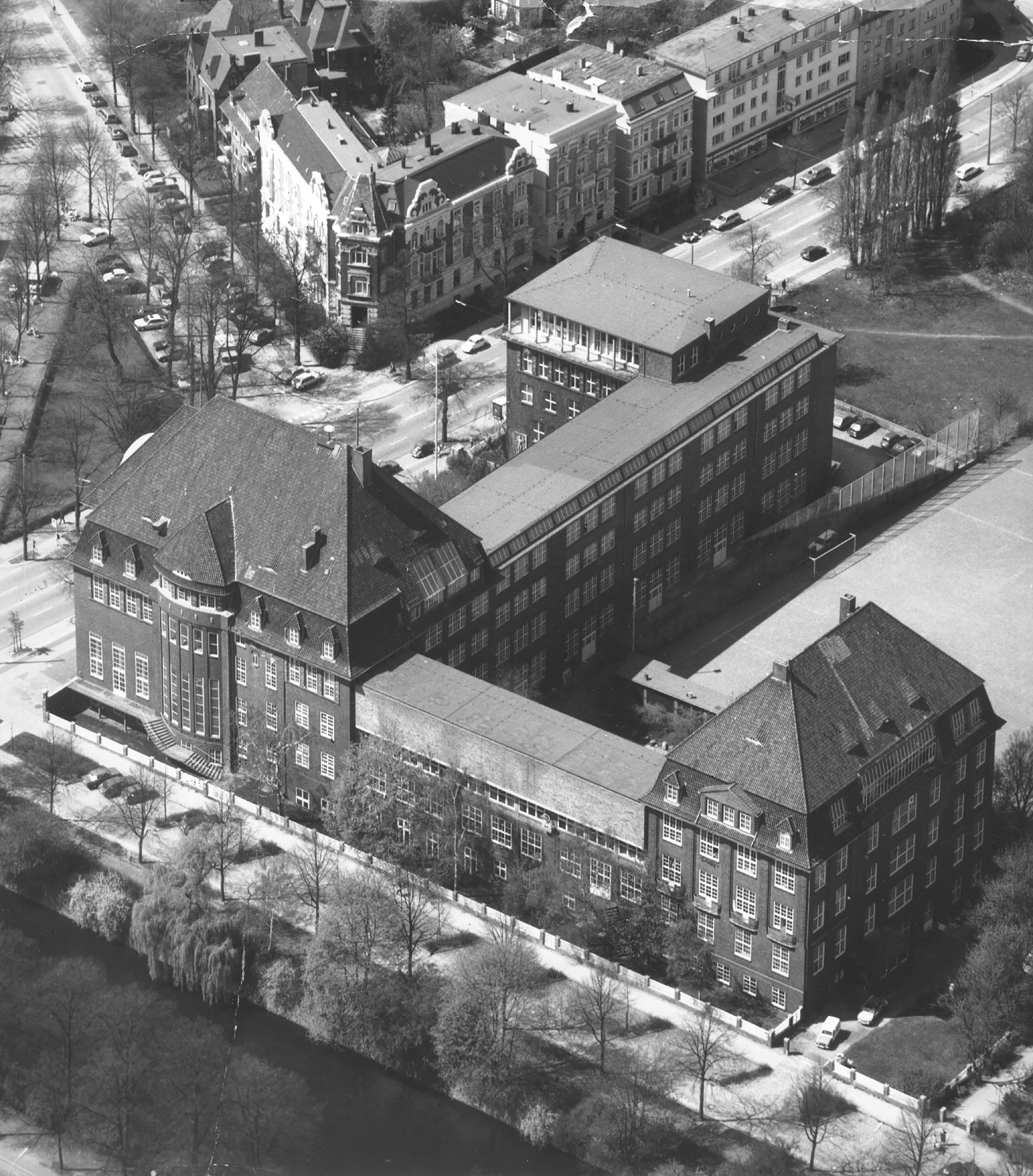 Historic black-and-white aerial photograph of a large brick building complex with steep gabled roofs; the ensemble is situated along a street and a body of water, surrounded by trees and neighboring residential buildings.