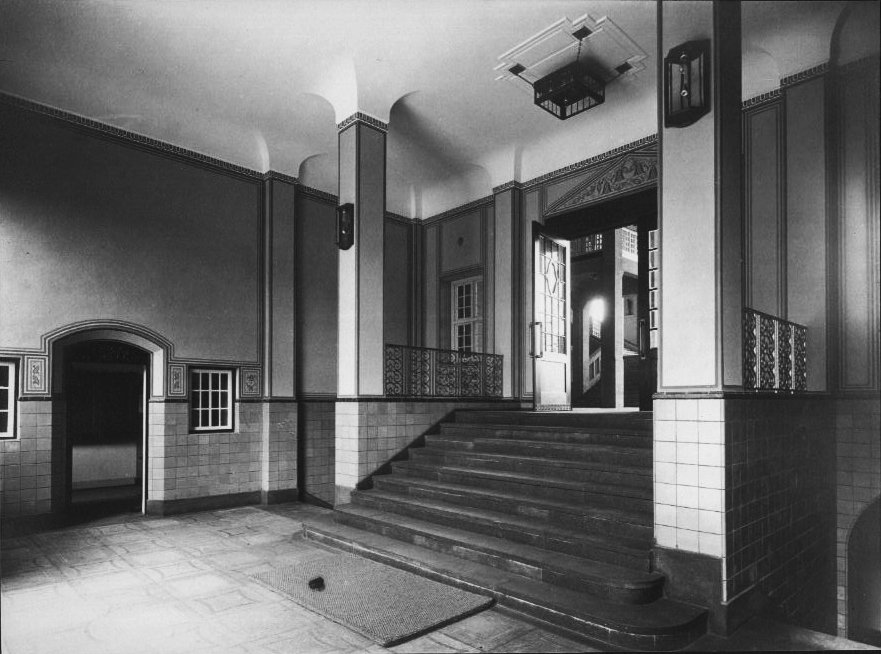 Historic black-and-white photograph of an entrance hall with a staircase, tall pillars, and decorative wall and ceiling details; an open door leads into another interior space.