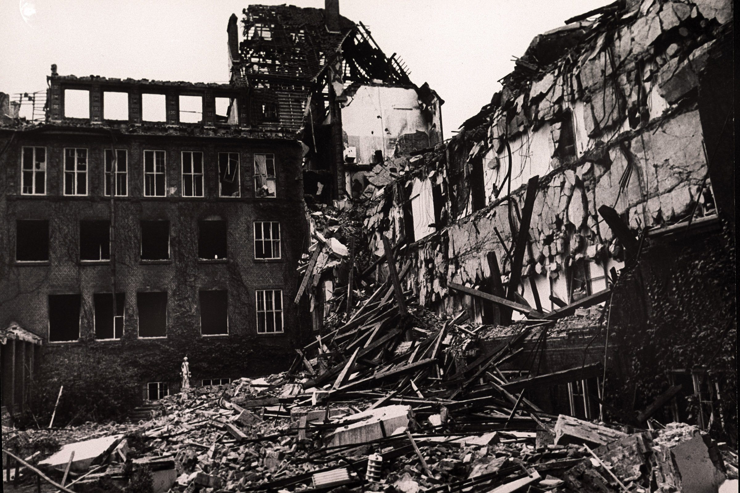Historic black-and-white photograph of a severely damaged building complex; collapsed roofs and walls, shattered windows, and large piles of rubble dominate the scene.