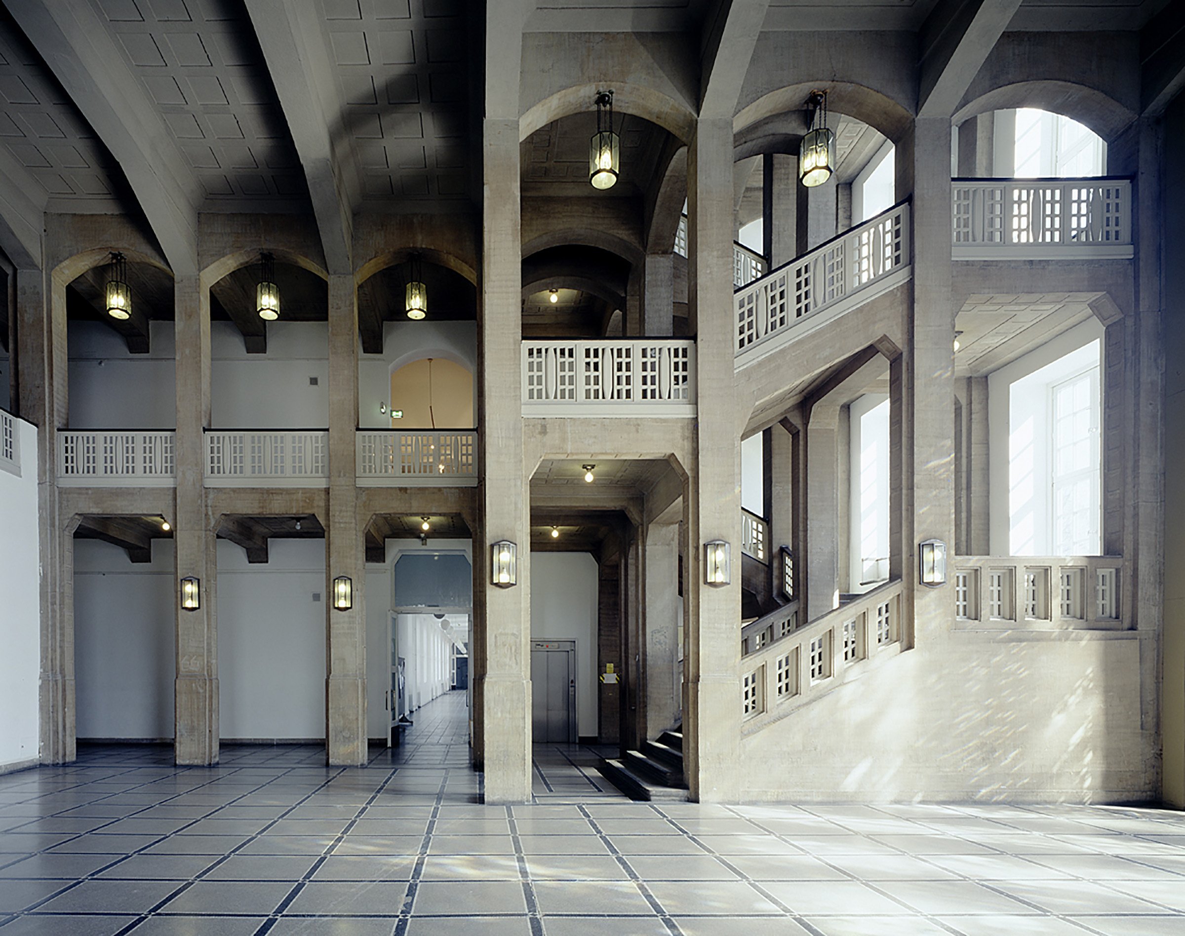 Spacious foyer with tall pillars, rounded arches, and a side staircase ascending along the wall; light stone surfaces, a geometric tiled floor, and hanging lamps define the historic interior.