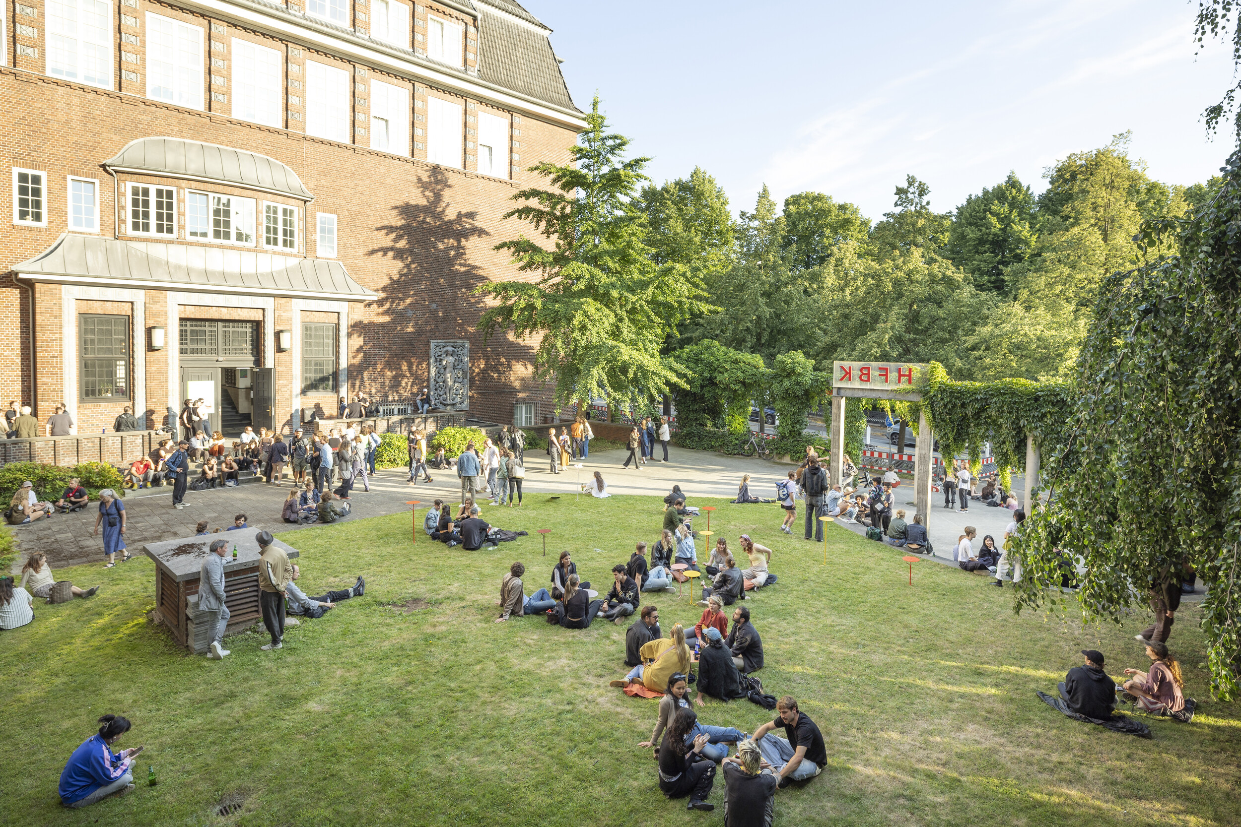 View of the green courtyard of a red brick building where many people are sitting on the lawn, standing, and talking; to the right is an ivy-covered pergola with the “HFBK” sign, with trees and summer light in the background.