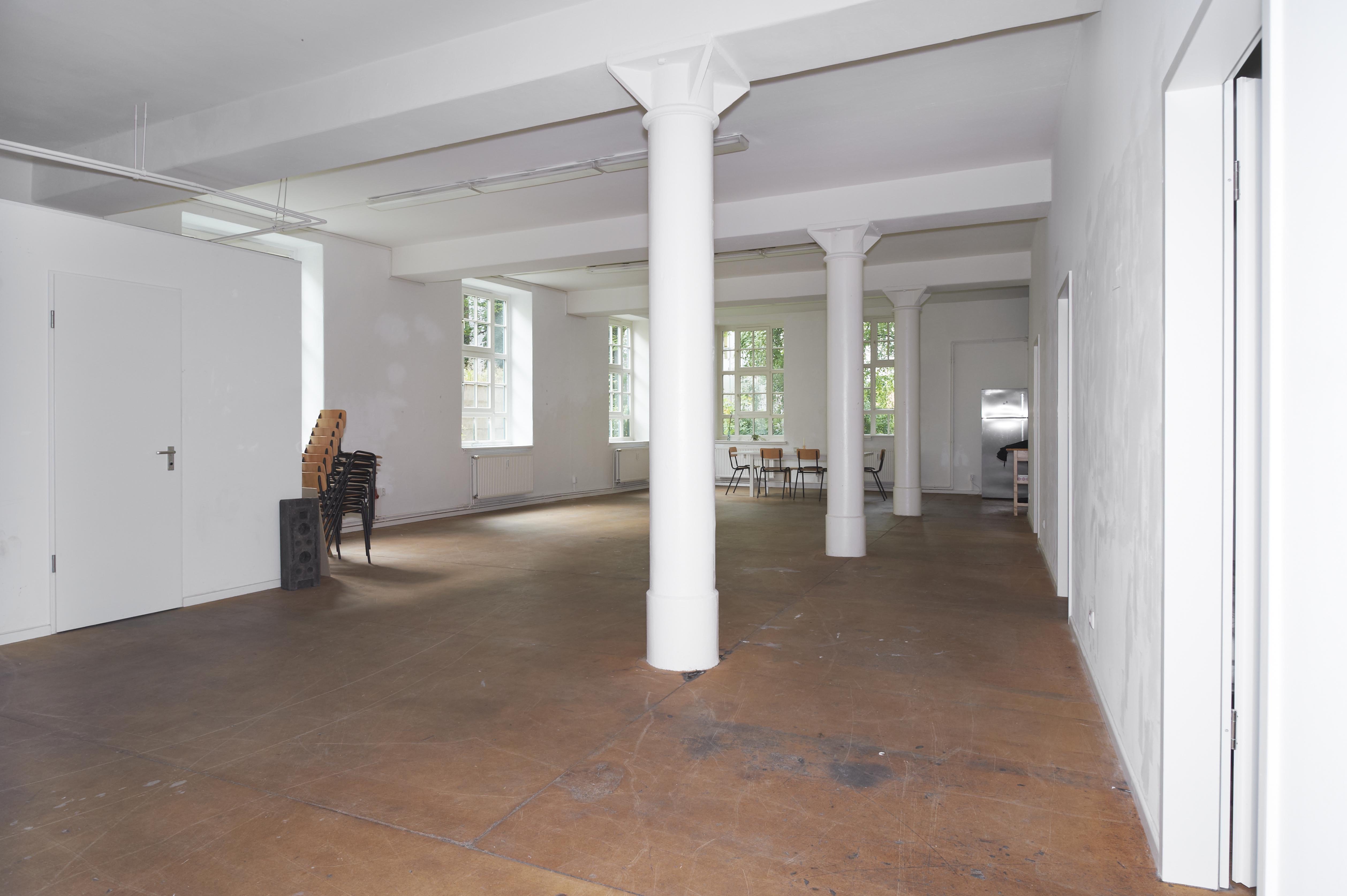 Bright, simple room with white walls and two large multi-pane windows. Radiators are located beneath the windows. The floor is brown and smooth.