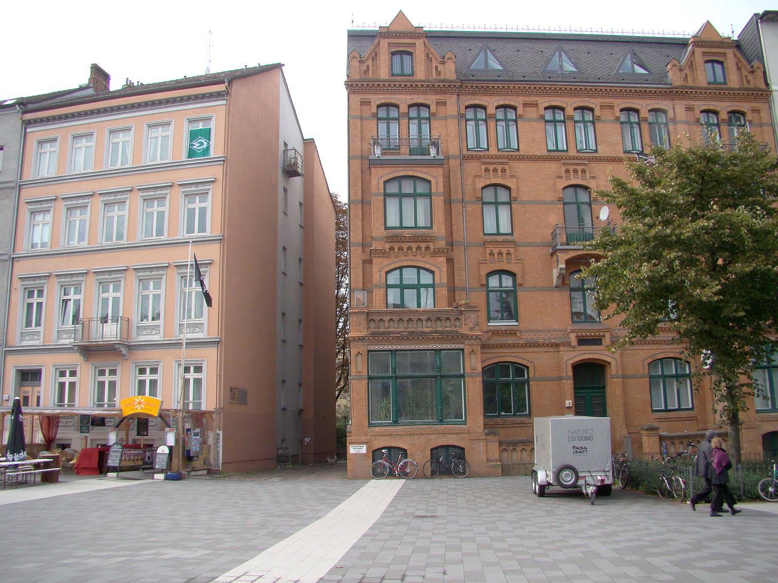 Two multi-story historic buildings on a paved square. On the left is a light-colored plastered building with white window frames and a small shop at ground level. On the right is a larger brick building with green-framed windows, balconies, and a roof with dormer windows. Bicycles and a small trailer are parked in front, and two people are walking on the right