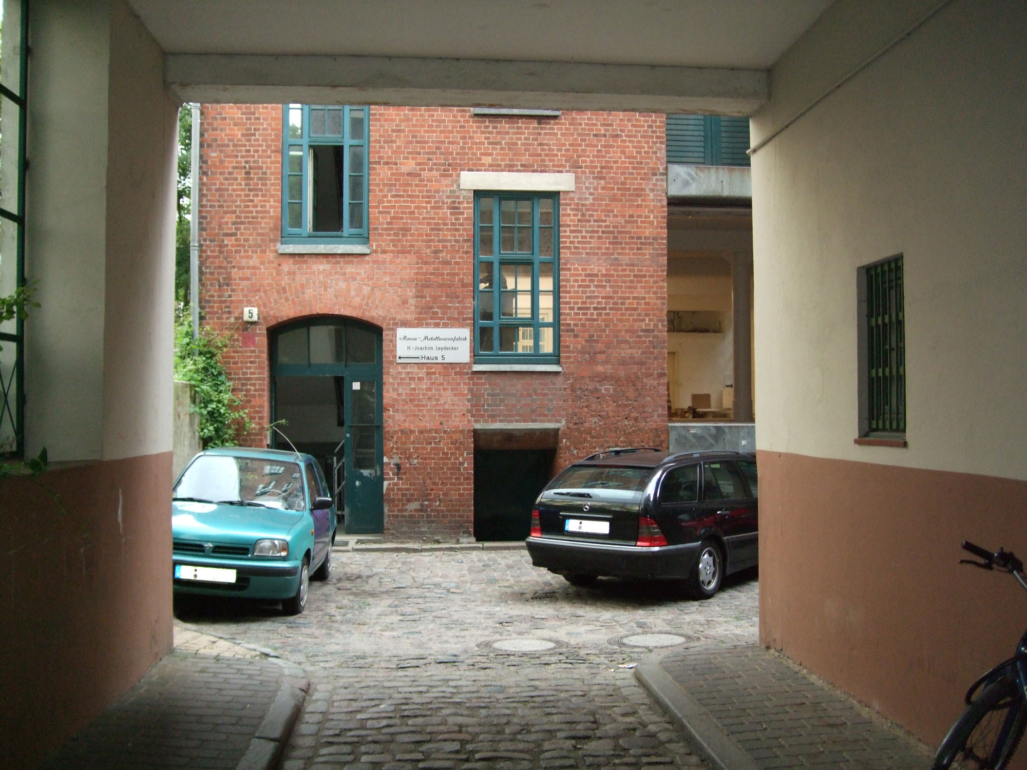 View through a passageway into a cobblestone courtyard. Opposite stands a red brick building with green-framed windows and the number 5 displayed near the entrance. Two cars are parked in the courtyard, a small turquoise vehicle on the left and a black station wagon on the right.