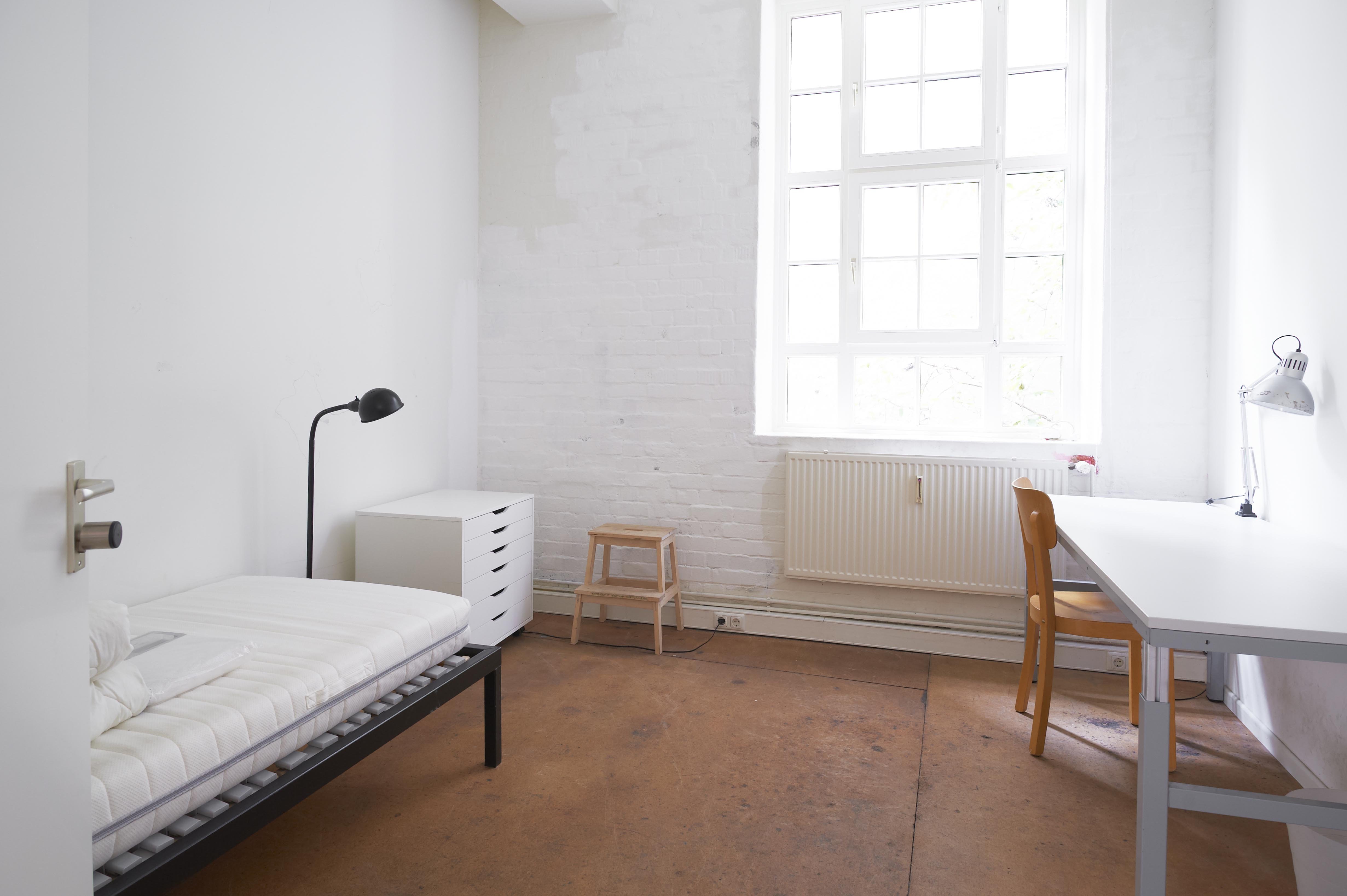 Bright, simple room with white walls and two large multi-pane windows. Radiators are mounted below the windows. In the corner there is a gray desk with a silver desk lamp on top, and a wastebasket underneath. The floor is smooth and brown.