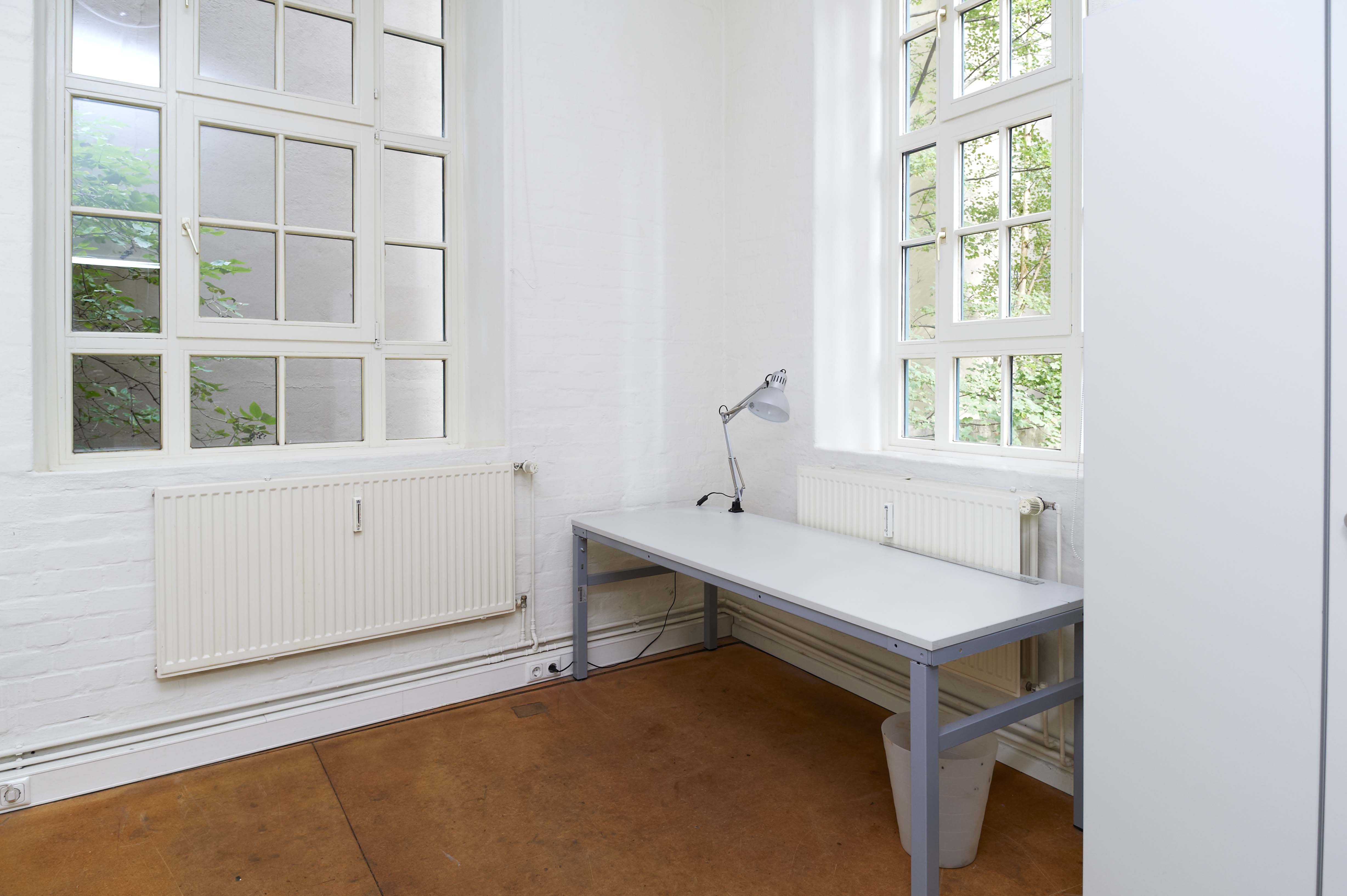Bright, simple room with white walls and two large multi-pane windows. Radiators are mounted below the windows. In the corner there is a gray desk with a silver desk lamp on top, and a wastebasket underneath. The floor is smooth and brown.