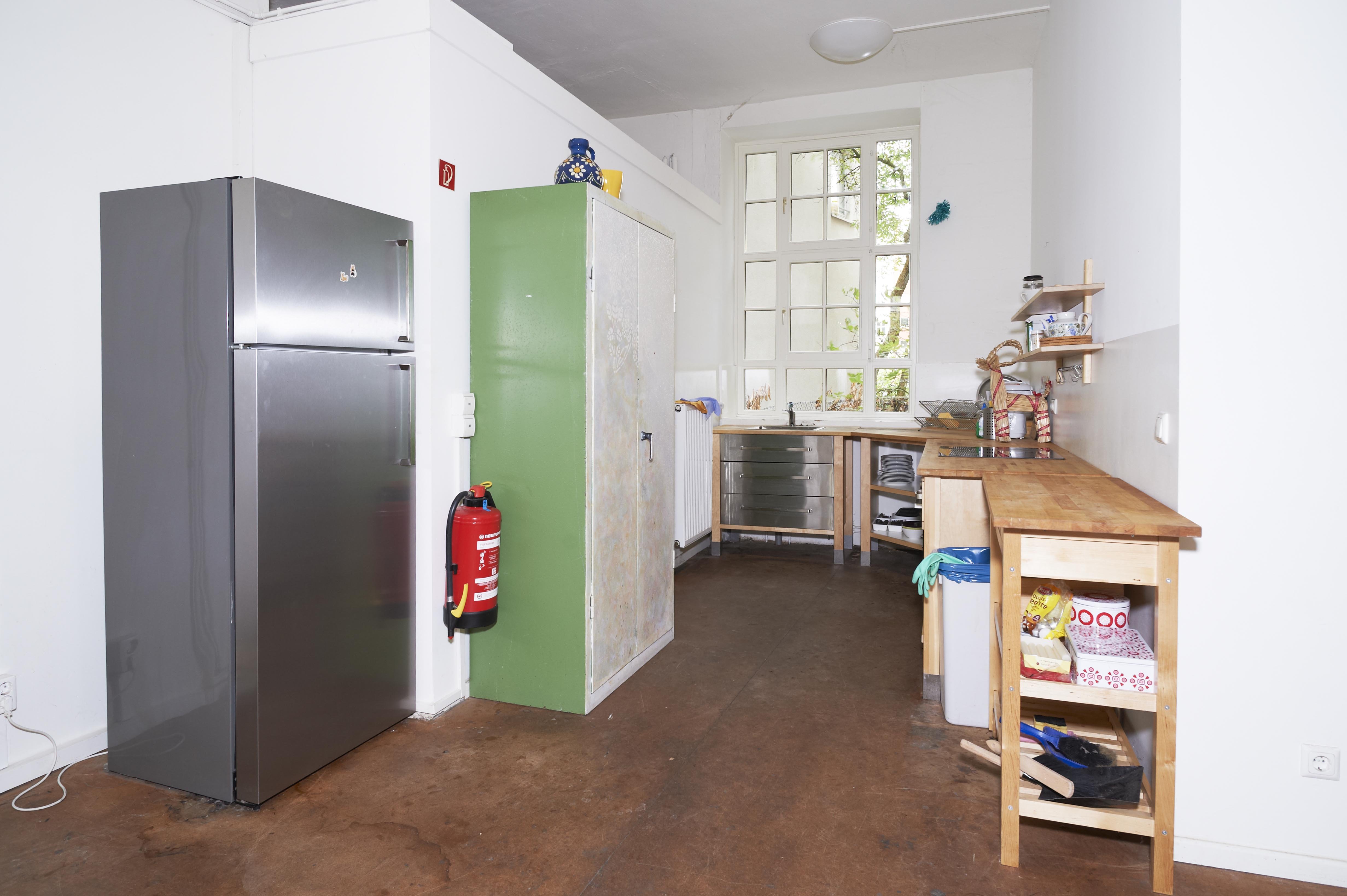 Small, bright kitchenette with white painted walls and a brown floor. On the left is a silver refrigerator next to a green cabinet with a fire extinguisher attached to its side. Beneath the window is a countertop with a sink and metal and wooden cabinets. On the right are additional wooden work surfaces and open shelves holding dishes and kitchen utensils.