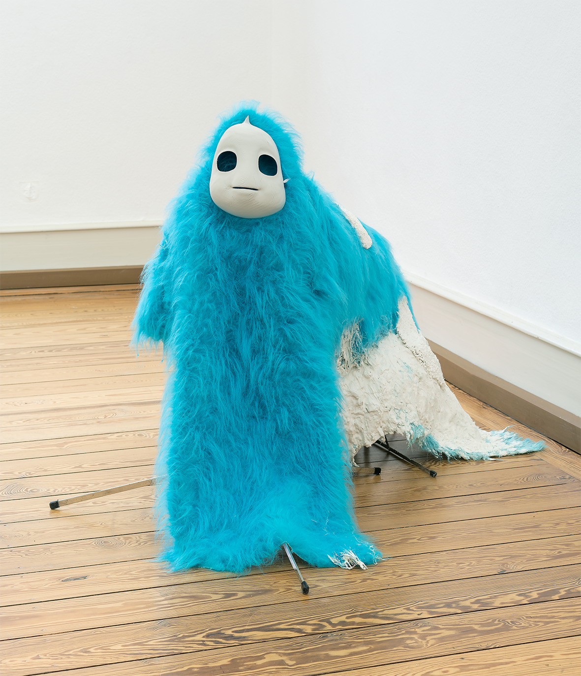 Skulptur oder Figur in einem Ausstellungsraum mit Holzboden und weißen Wänden. Die Figur ist mit langem, türkisfarbenem Kunstfell bedeckt und trägt eine glatte, weiße Maske mit großen, dunklen Augenöffnungen. Hinter ihr liegt ein teilweise weißes, textilartiges Element auf dem Boden.