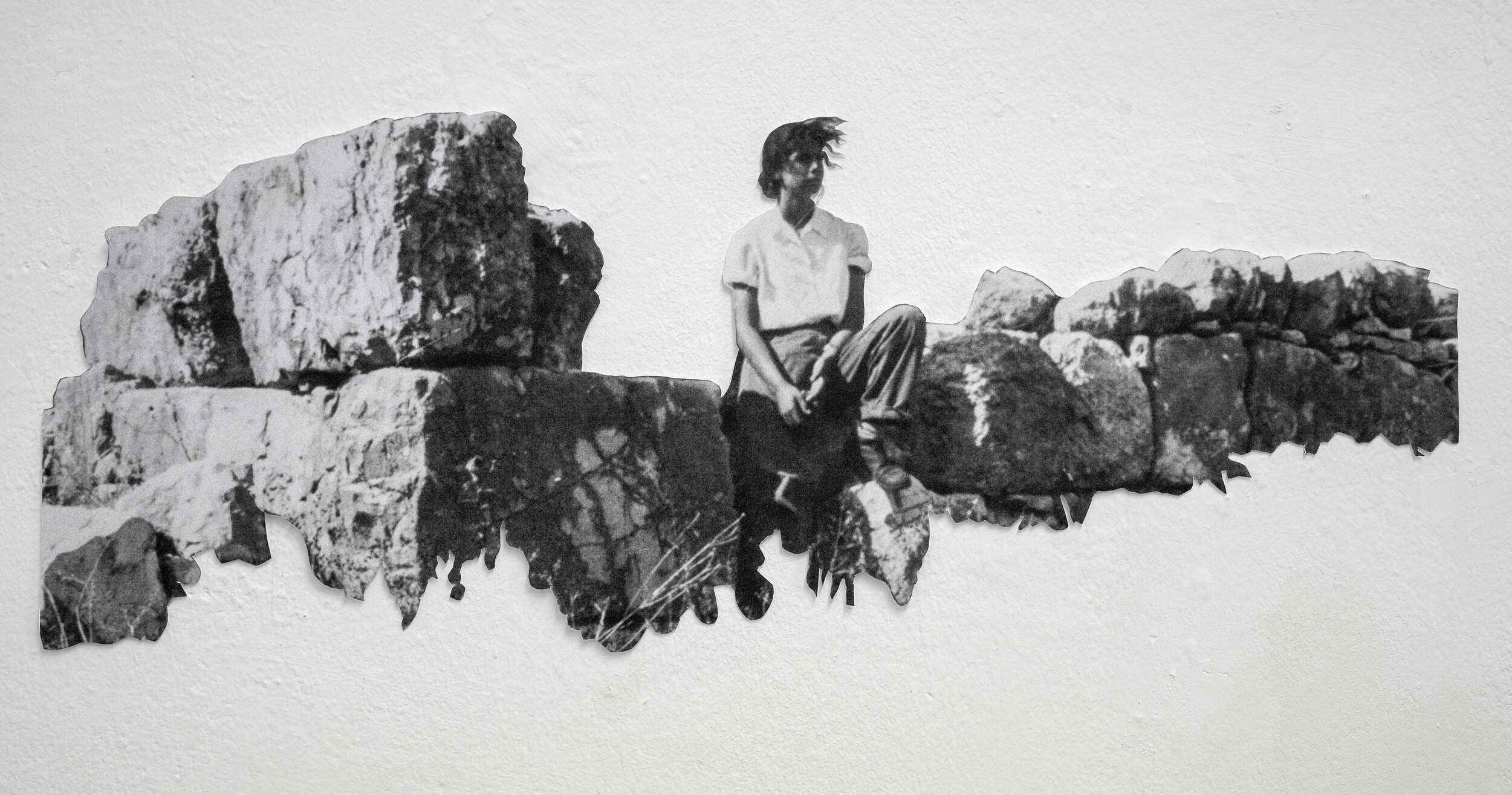 Schwarz-weiße fotografische Arbeit an einer weißen Wand. Zu sehen ist eine ausgeschnittene Darstellung einer Person, die auf einer Steinmauer zwischen großen Felsblöcken sitzt. Die unregelmäßigen Kanten des Bildes sind sichtbar und betonen den collageartigen Charakter.