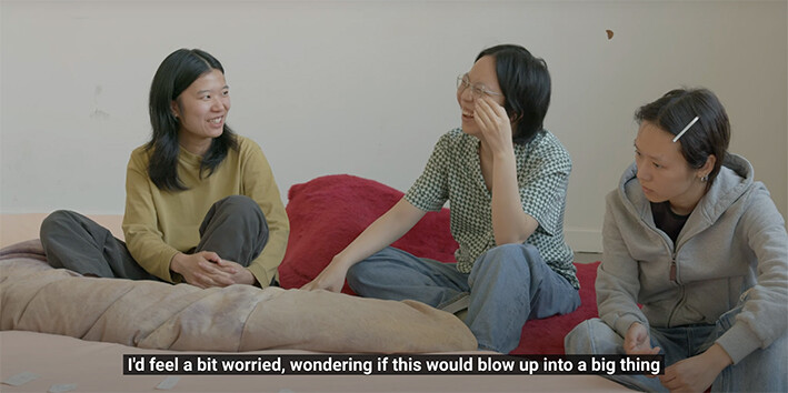 Drei Personen sitzen auf Kissen auf dem Boden in einem hellen Raum und unterhalten sich. Zwei von ihnen lachen, während die dritte aufmerksam zuhört. Unten im Bild ist ein englischer Untertitel eingeblendet: „I’d feel a bit worried, wondering if this would blow up into a big thing“.
