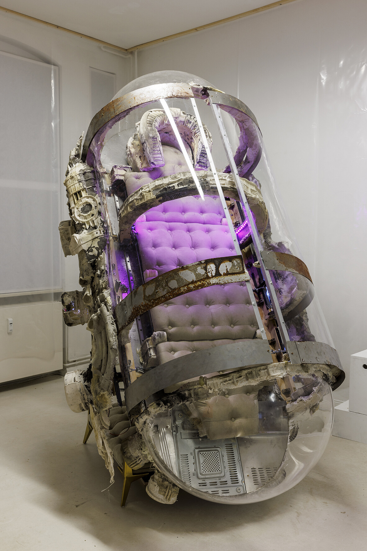 Großformatige, kapselartige Skulptur in einem weißen Ausstellungsraum. Die Konstruktion besteht aus transparentem Kunststoff und Metallbändern mit rostigen Spuren. Im Inneren befinden sich übereinander angeordnete, gepolsterte Sitze in Rosa- und Beigetönen, die von violettem Licht beleuchtet werden. Technische Bauteile und Kabel sind sichtbar in die Struktur integriert.