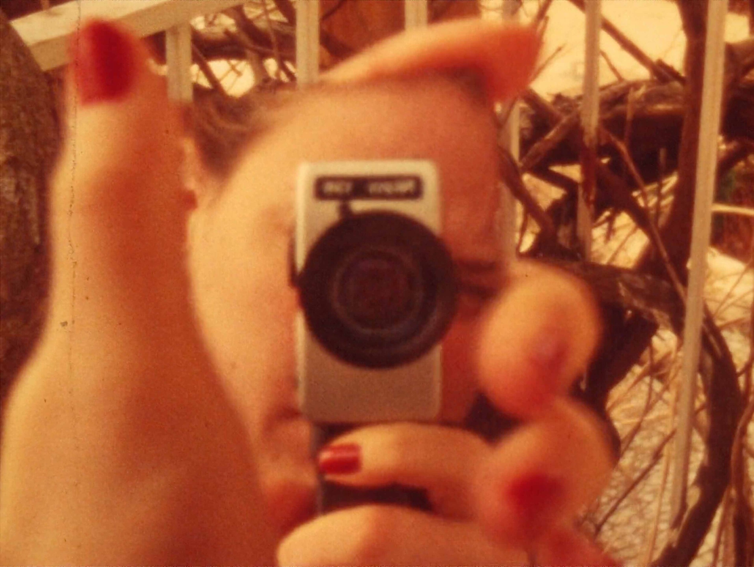 Nahaufnahme einer Person, die eine kleine Kamera direkt vor ihr Gesicht hält und fotografiert. Die Kamera verdeckt die Augenpartie. Die Fingernägel sind rot lackiert. Das Bild hat einen warmen, leicht unscharfen Vintage-Look.