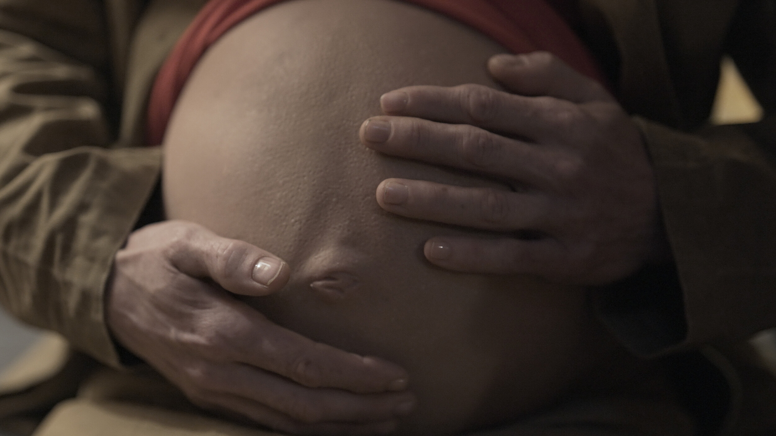 Nahaufnahme eines schwangeren Bauchs. Zwei Hände umfassen und stützen den Bauch von beiden Seiten. Die Haut ist deutlich sichtbar, und am oberen Rand ist ein Stück roter Kleidung zu erkennen. Die Szene ist in warmem, gedämpftem Licht aufgenommen.