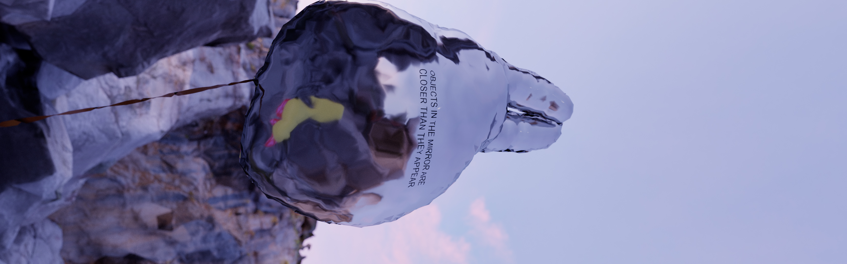 Detail of a digital scene: A reflective floating form hangs among rocks in a barren landscape. The phrase “OBJECTS IN THE MIRROR ARE CLOSER THAN THEY APPEAR” is written on its surface.