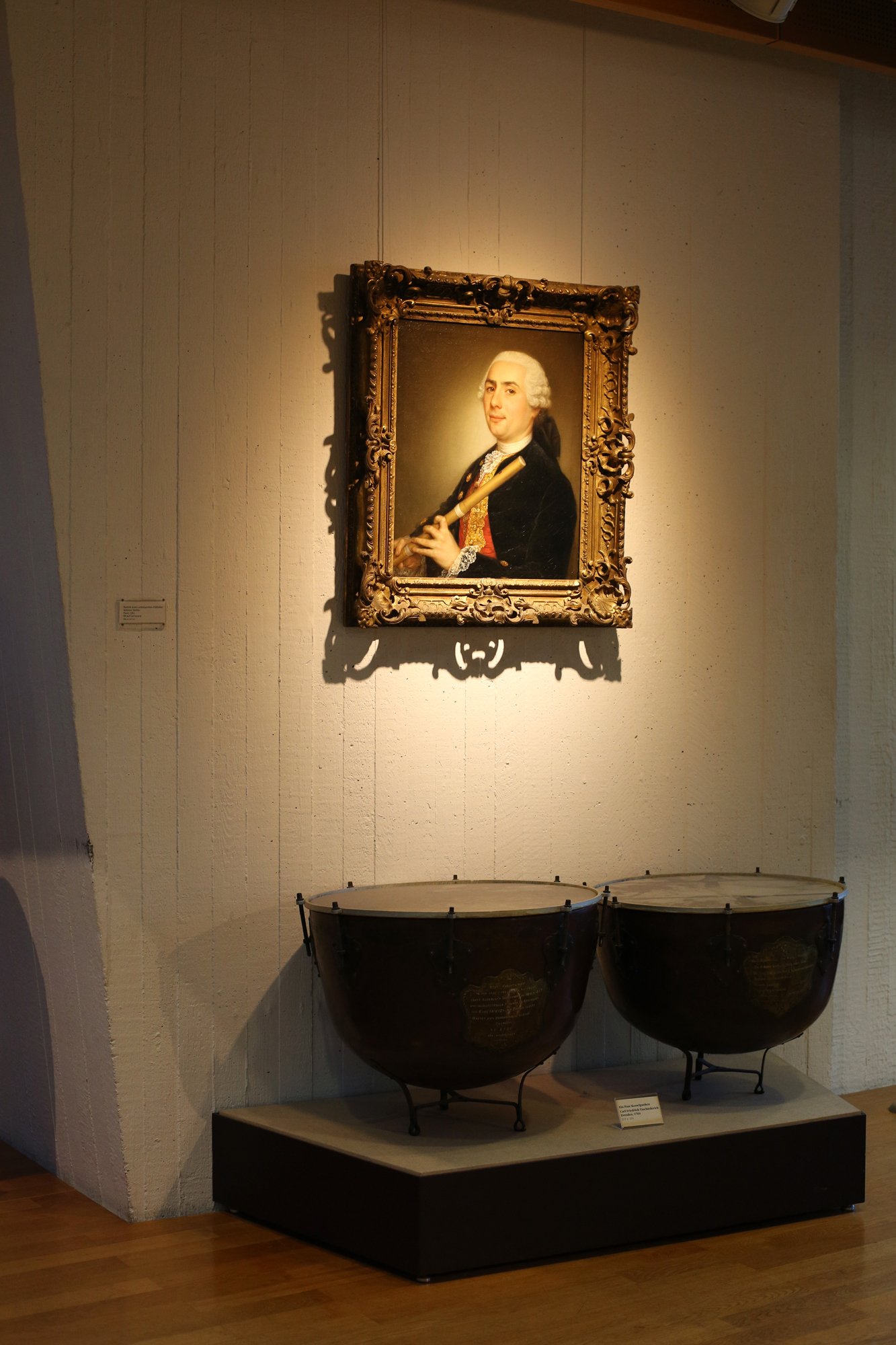 Eighteenth-century portrait of a man holding a flute on the wall, with two historic kettledrums displayed below in a museum.