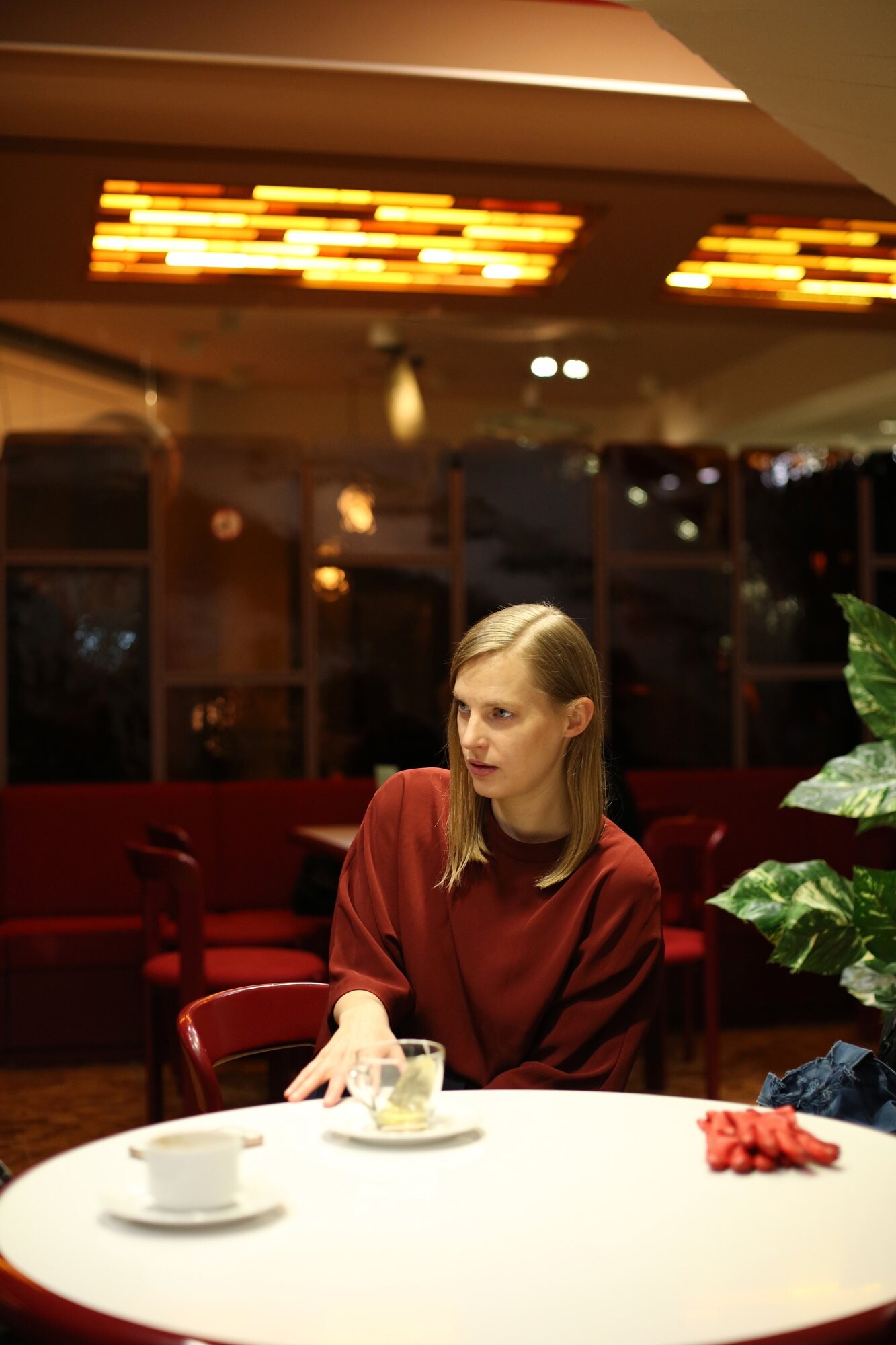 A person sitting at a café table with a cup of tea, in a warmly lit room with red seating and plants.