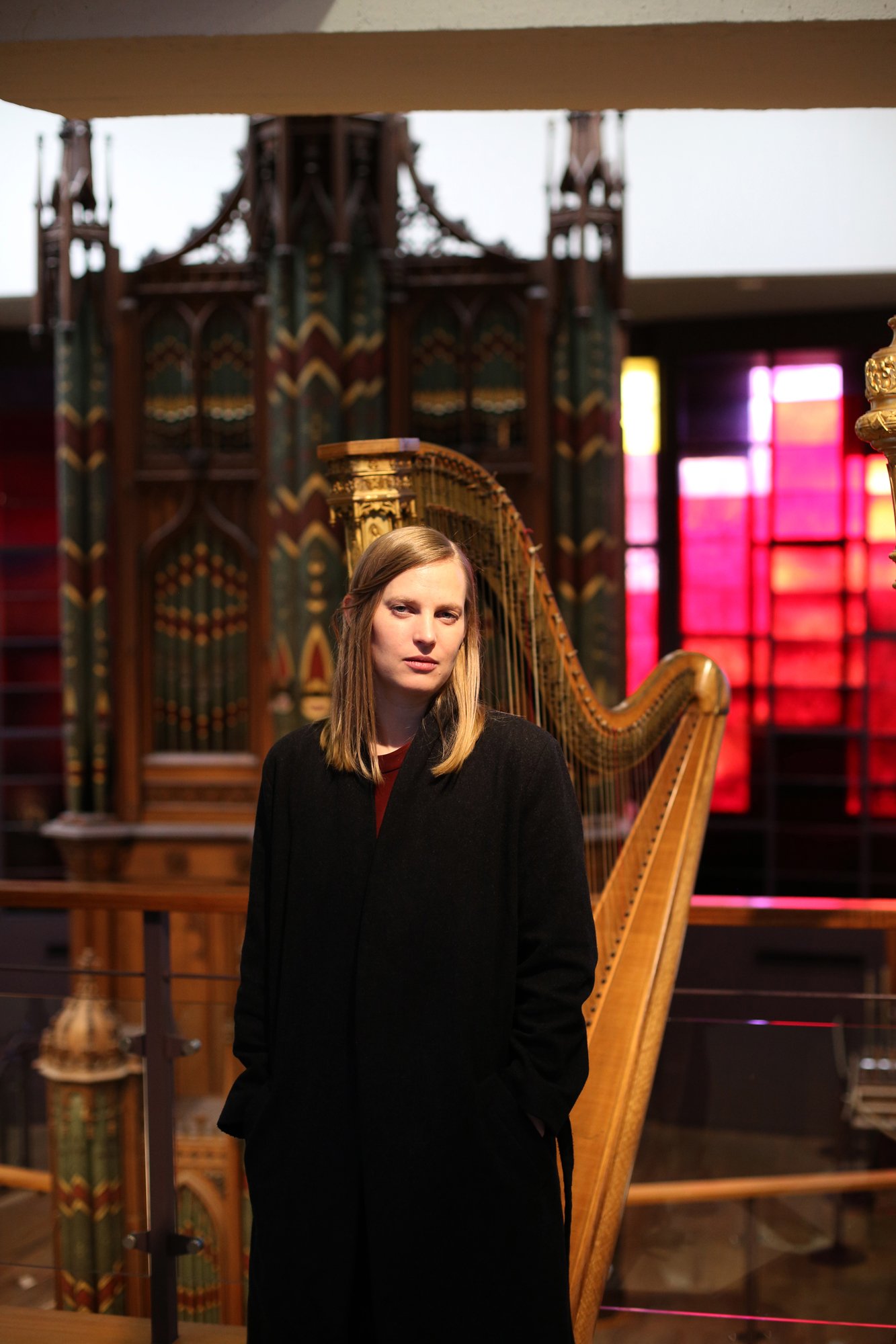 A person with long blonde hair standing in front of a decorative harp in a museum, with colorful stained-glass windows in the background.
