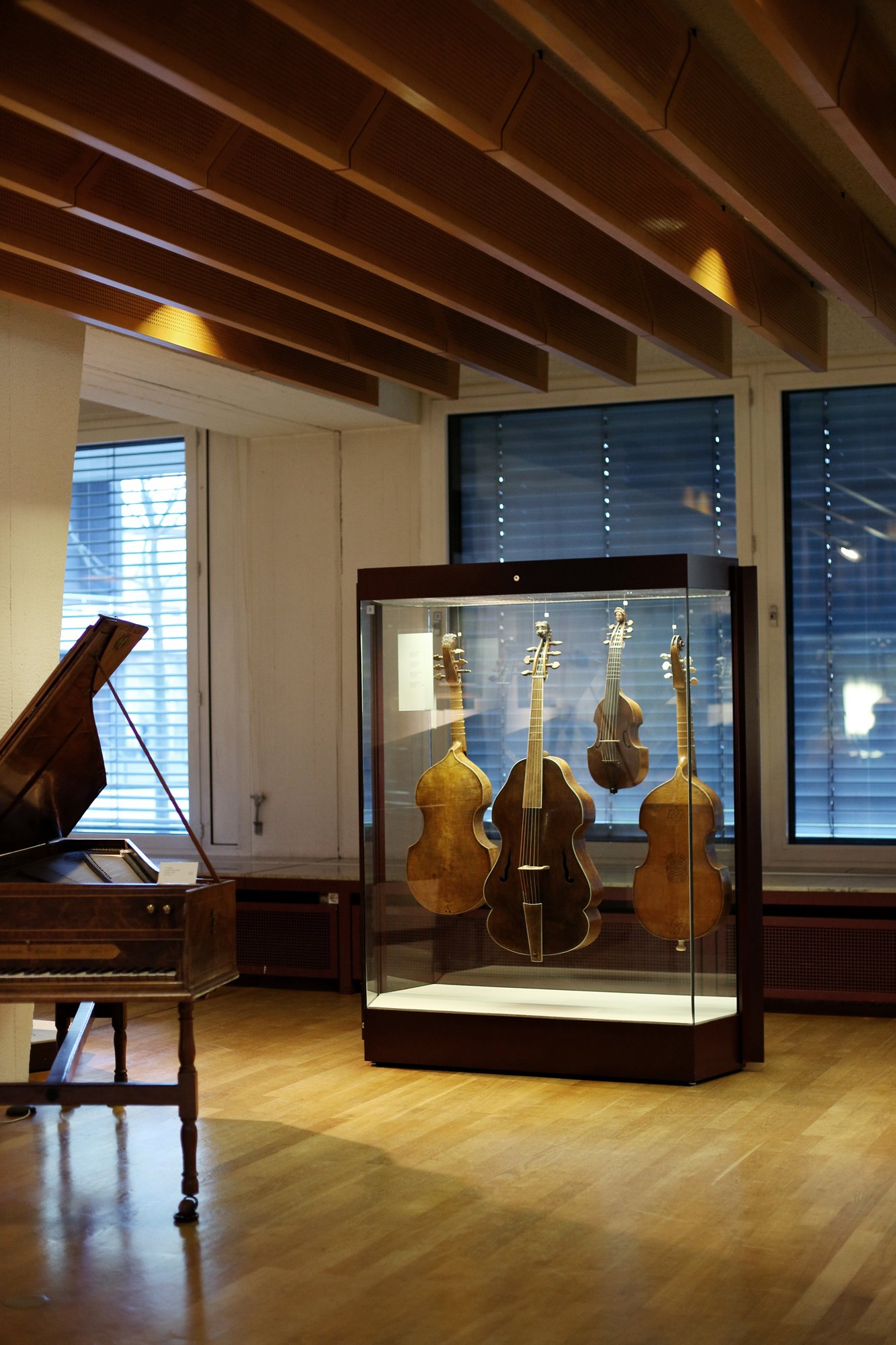 Historic string instruments displayed in a glass case in a museum, with an old keyboard instrument beside it in a wooden-ceiling exhibition room.