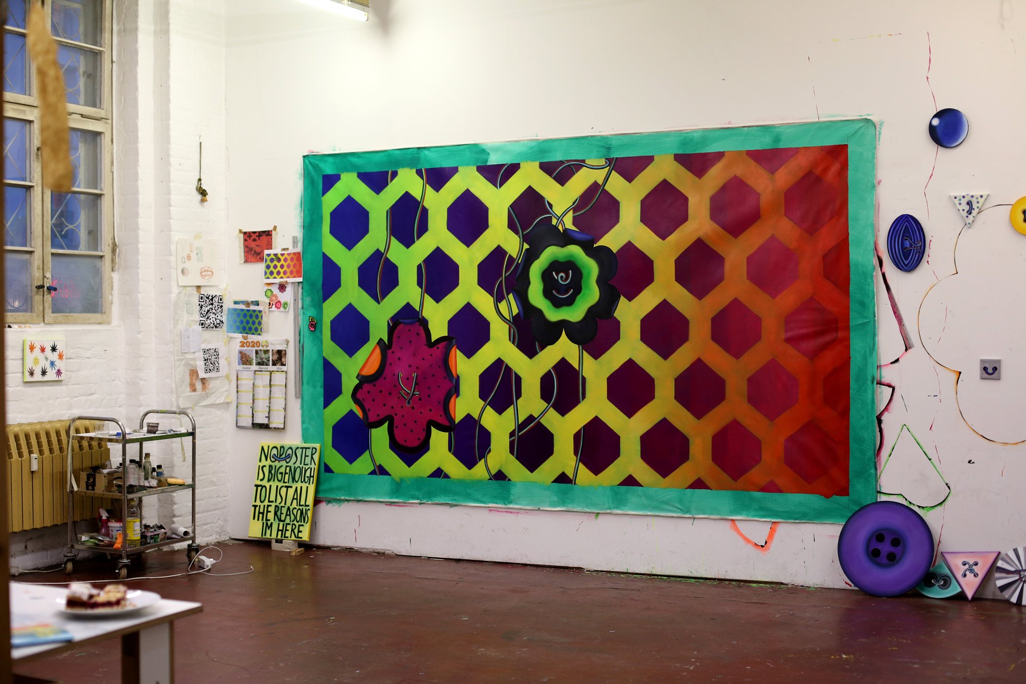 A studio space with a large, colorful wall piece featuring a hexagonal grid in yellow, green, and violet, along with two stylized flower shapes. Smaller artworks and a shelf with materials are positioned nearby.