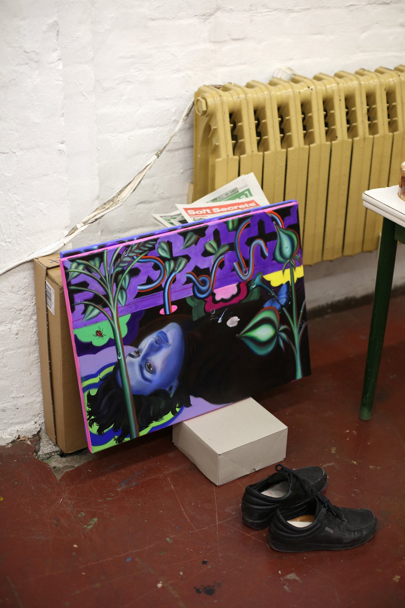 A colorful painting of a reclining blue-toned figure with plant motifs rests against a yellow radiator. Next to it are cardboard boxes, a newspaper, and a pair of black shoes.