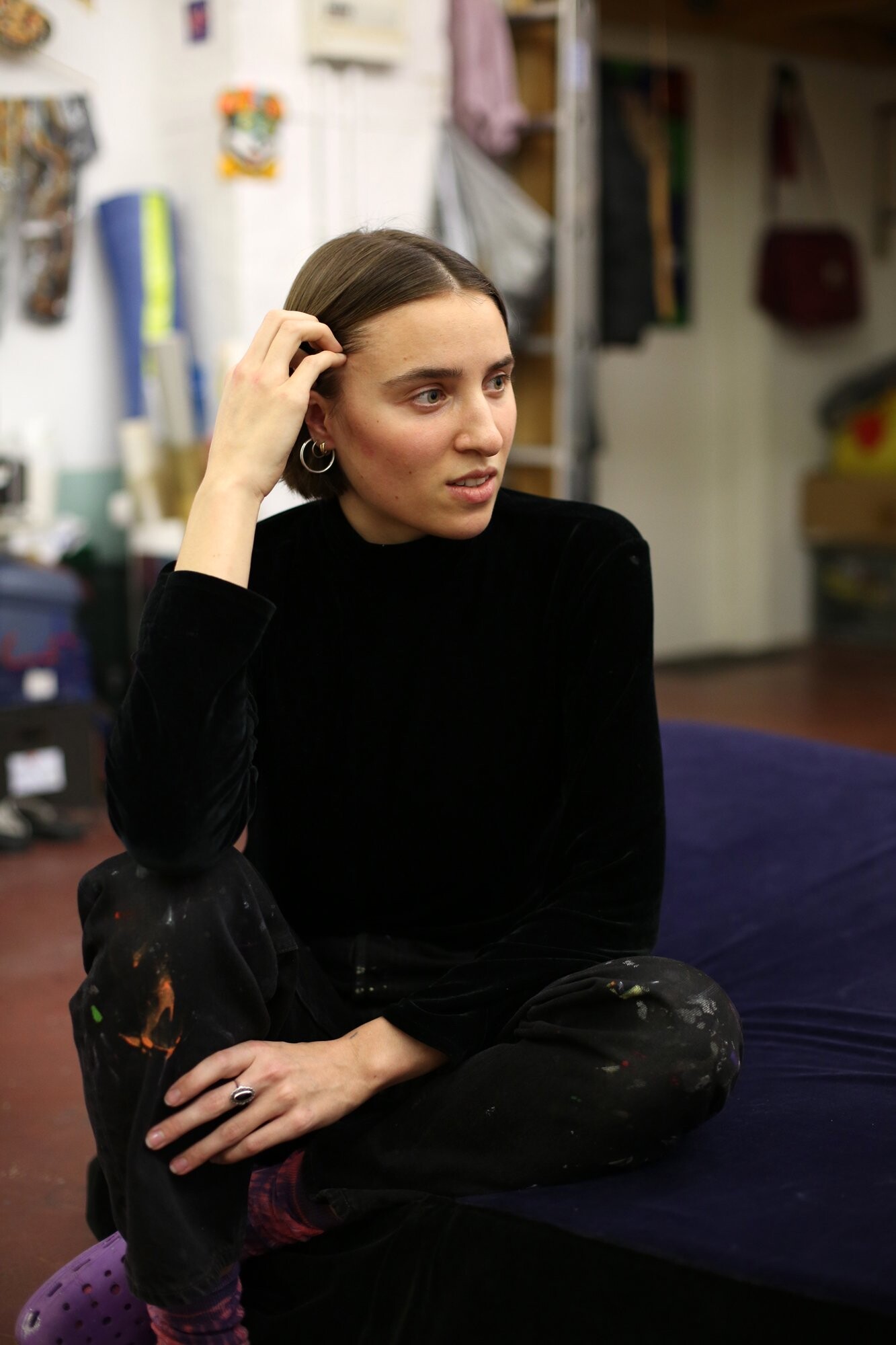 A person wearing paint-stained clothing sits on a dark cushion in a studio, touching their head with one hand.