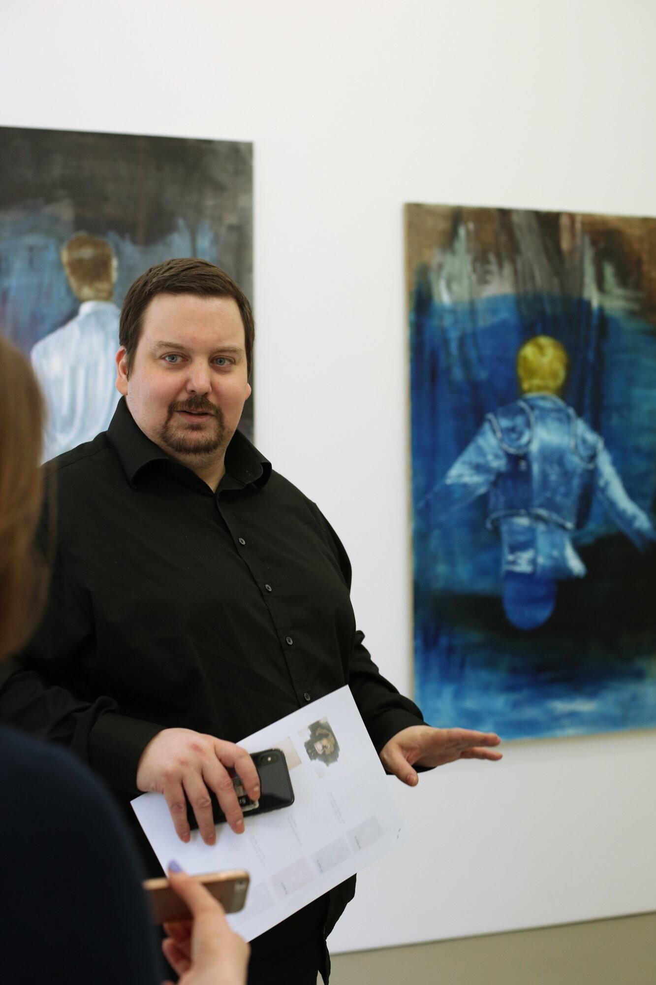 A person in a black shirt speaks inside a gallery, standing in front of two large paintings featuring figures seen from behind in blue and brown tones.