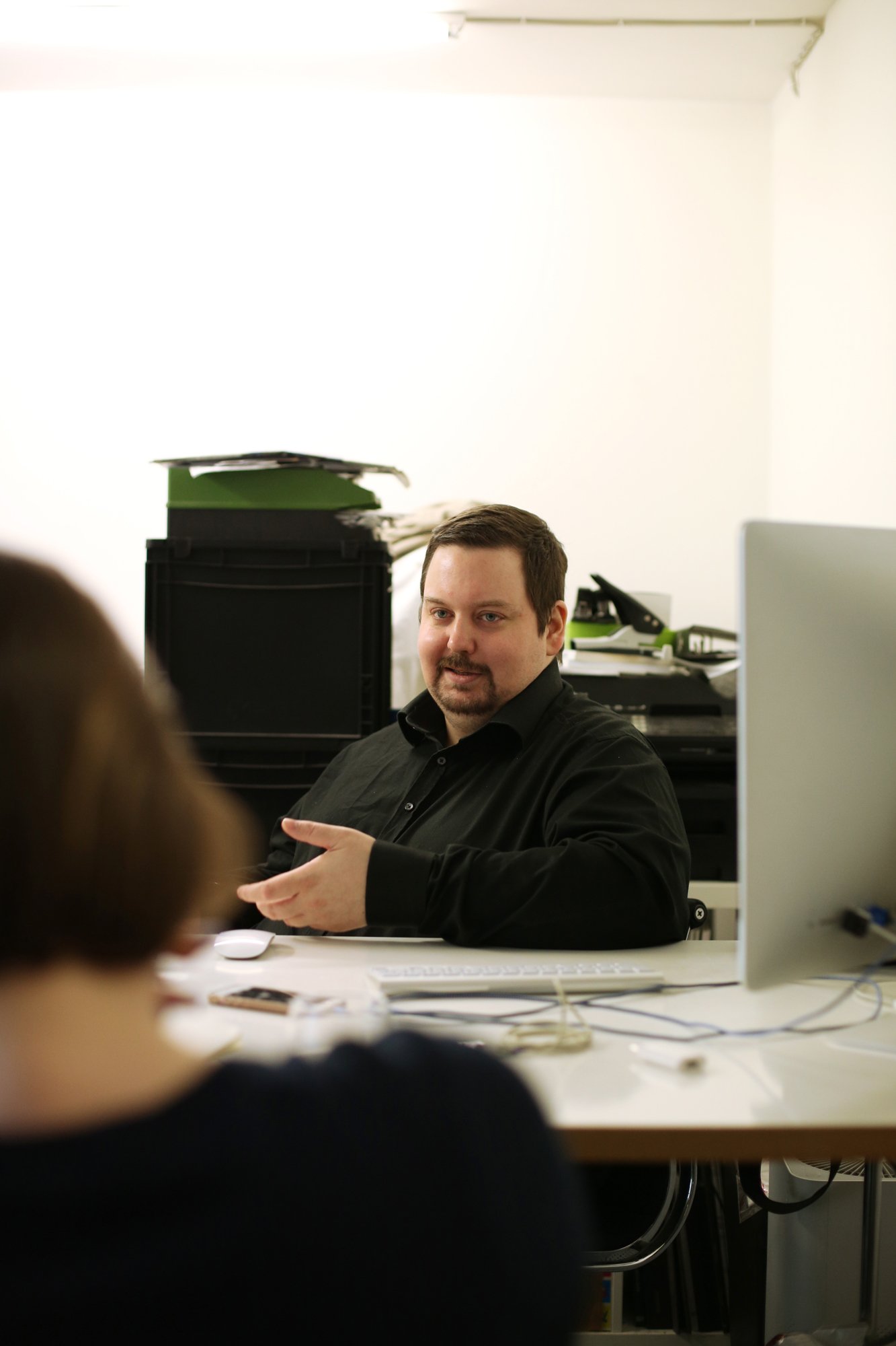 A person sits at a desk in a studio or office, gesturing while speaking; a computer and various documents are on the table.