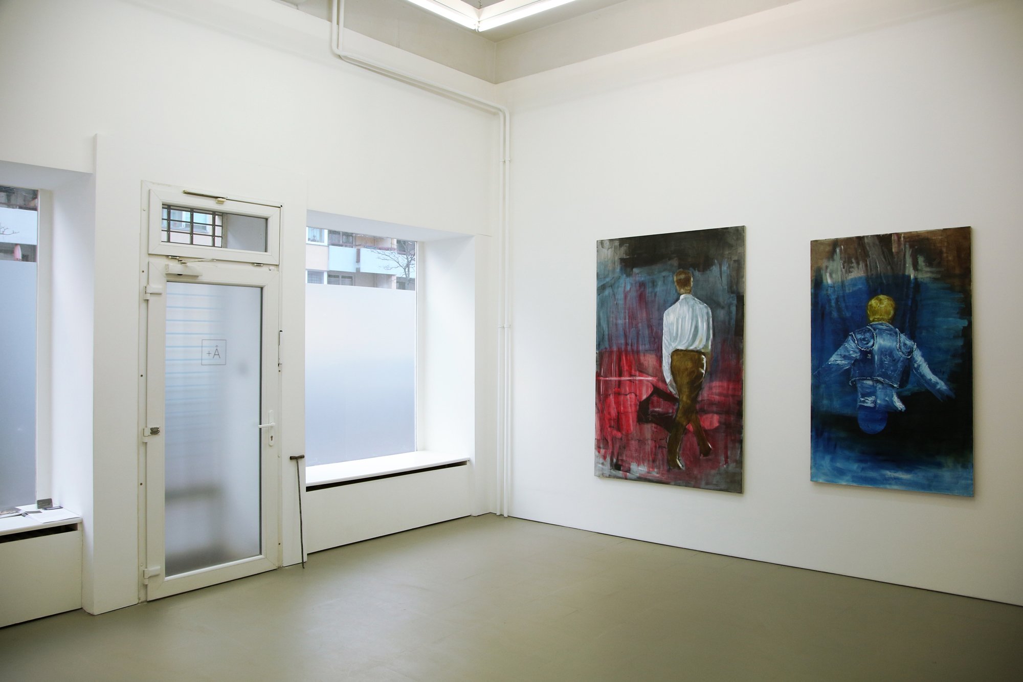 Bright gallery space with two large paintings on the wall and a glass entrance door displaying the “Å+” logo.
