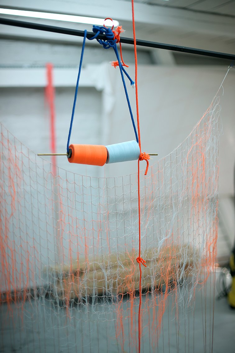 Orange and light blue spools of thread hang from cords attached to a metal rod above a loosely woven transparent net. Workshop details appear blurred in the background.