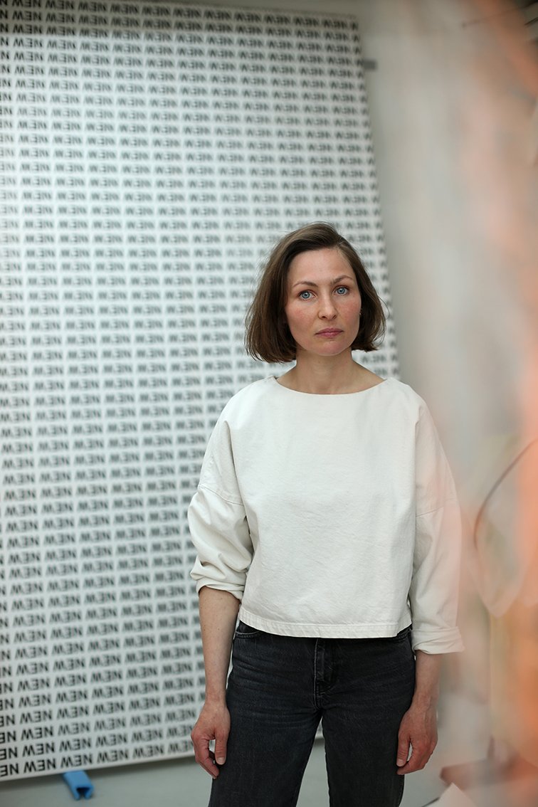 A person with short hair wearing a white top stands in a room in front of a wall densely covered with the word “MEN” in black typography.