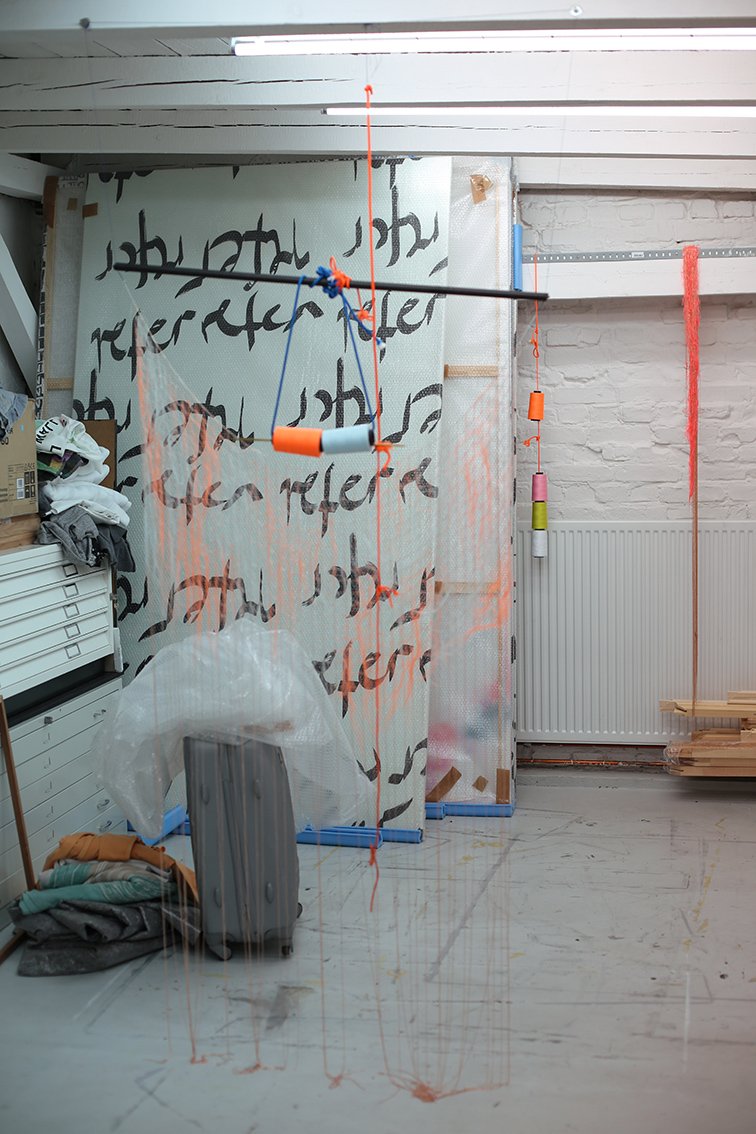 A studio space featuring suspended cords, spools of thread, and netting. In the background there is a large surface covered in black lettering, with fabric, wood, and packaging material stacked nearby.