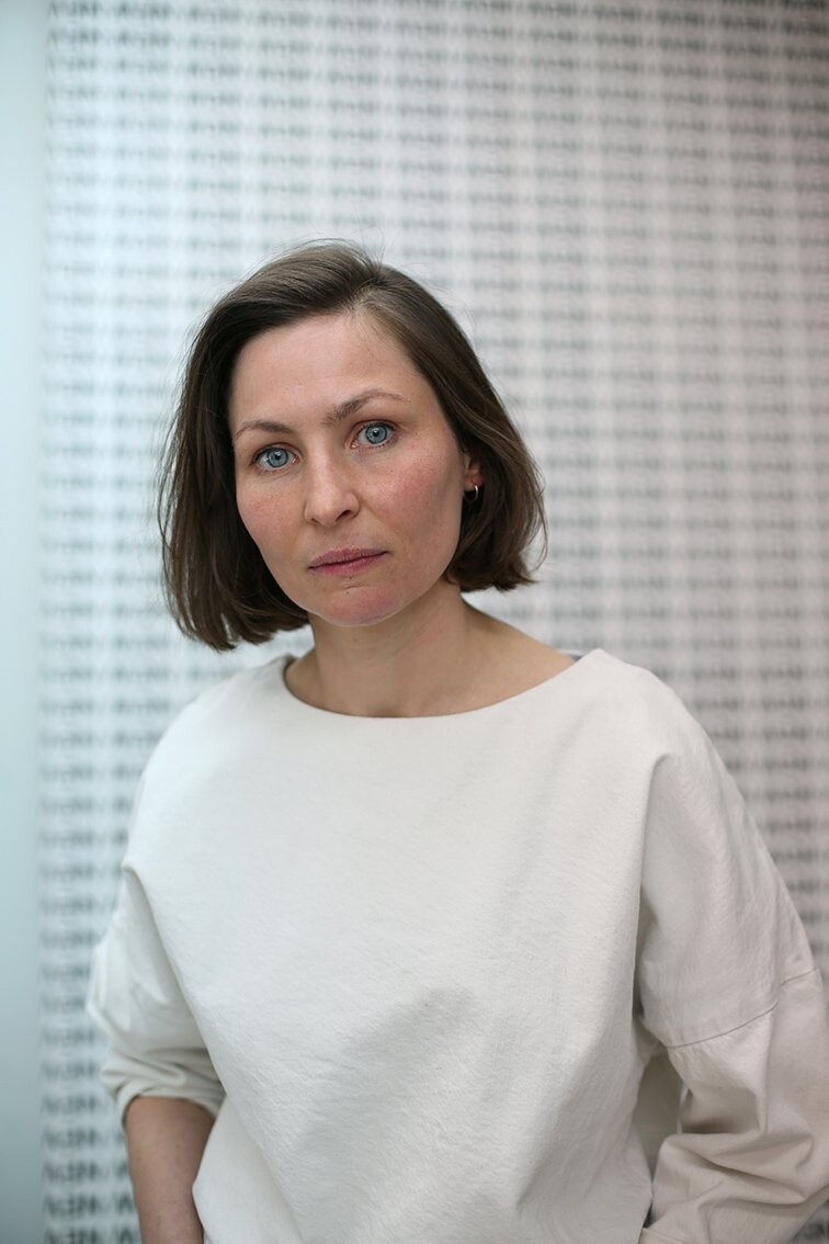 A person wearing a light-colored top, visible from the waist up, stands in front of a wall covered with repeated black lettering. The background is softly out of focus.