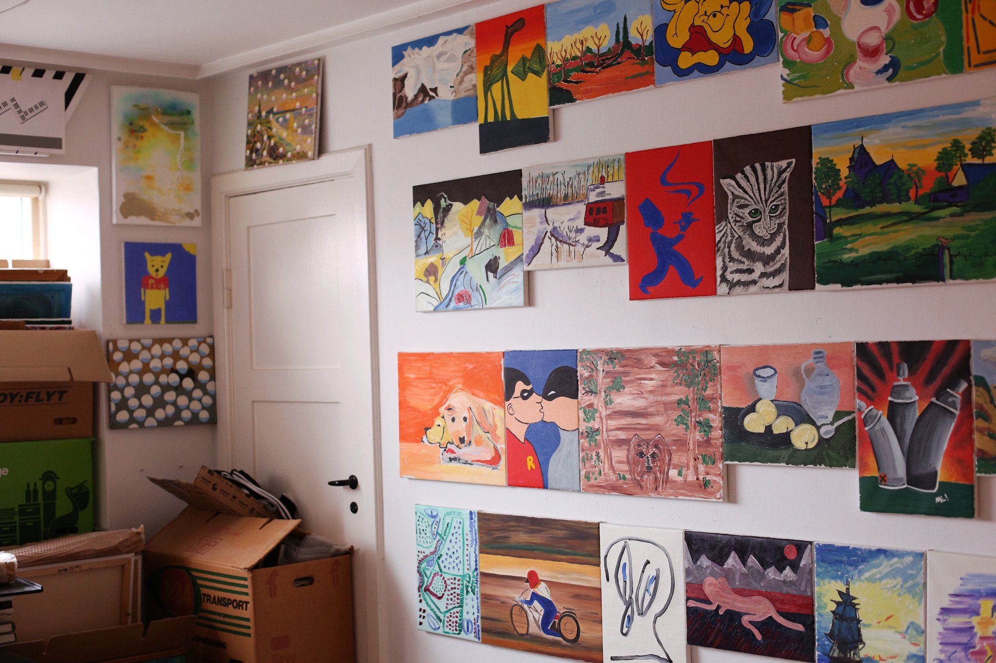 A room with densely hung small paintings depicting various motifs such as animals, landscapes, and figures. Cardboard boxes and packing material are stacked below.