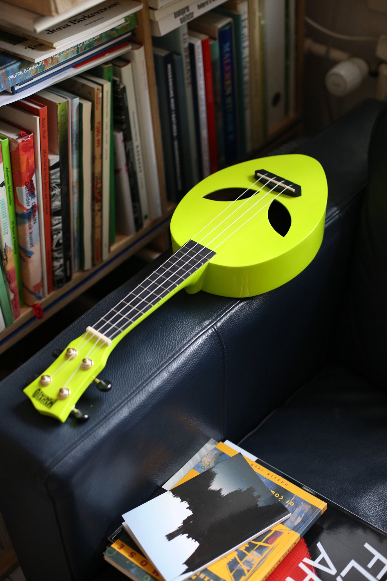 A neon yellow ukulele rests on the arm of a dark leather chair next to a stack of books and magazines.