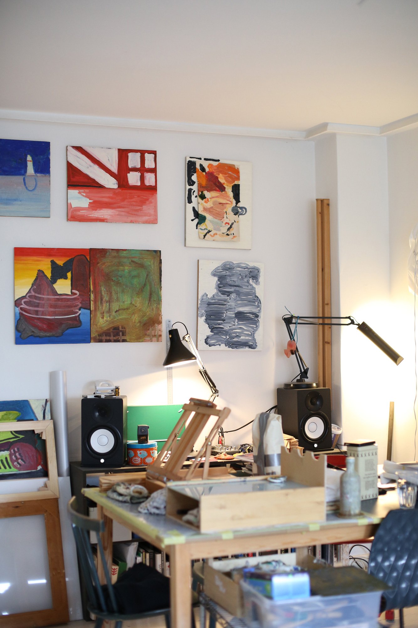 A workspace in an art studio with several small colorful paintings on the wall. Brushes, tools, and speakers sit on the table, lit by two desk lamps.