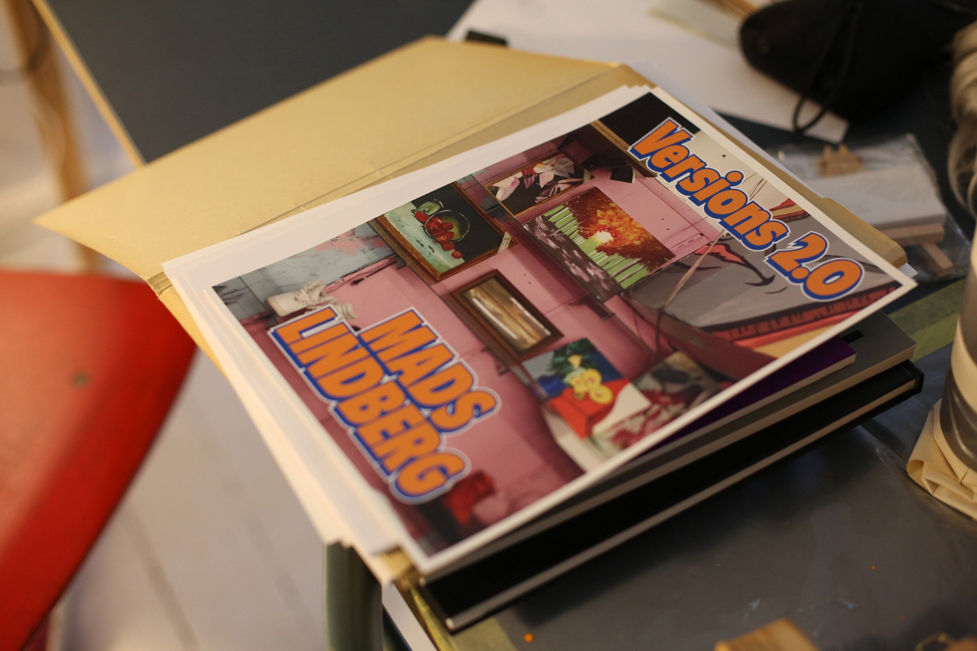 A pile of printed material lies on a table, topped with a booklet titled “Versions 2.0” and the name “MADS LINDBERG,” featuring a room filled with paintings on the cover.