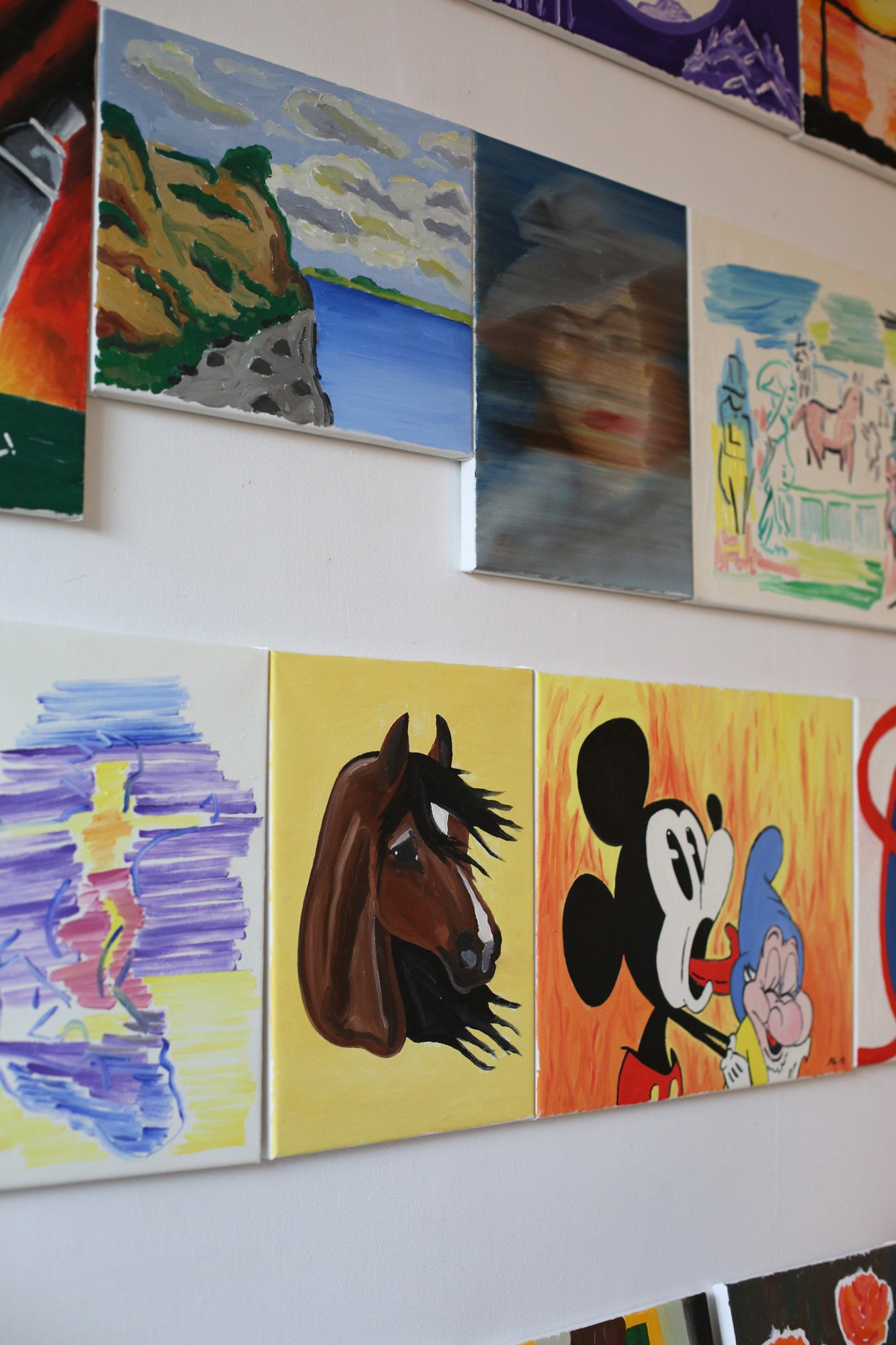 Close-up of several paintings on a wall, including landscapes, a horse depiction, and a character resembling a cartoon style.