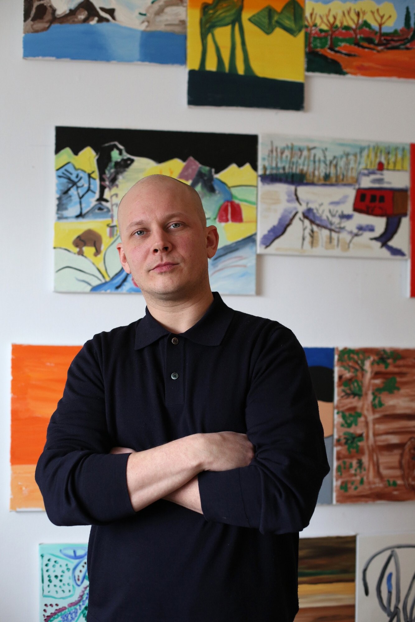 A person stands with their arms crossed in front of a wall covered in colorful small paintings depicting landscapes, figures, and animals.