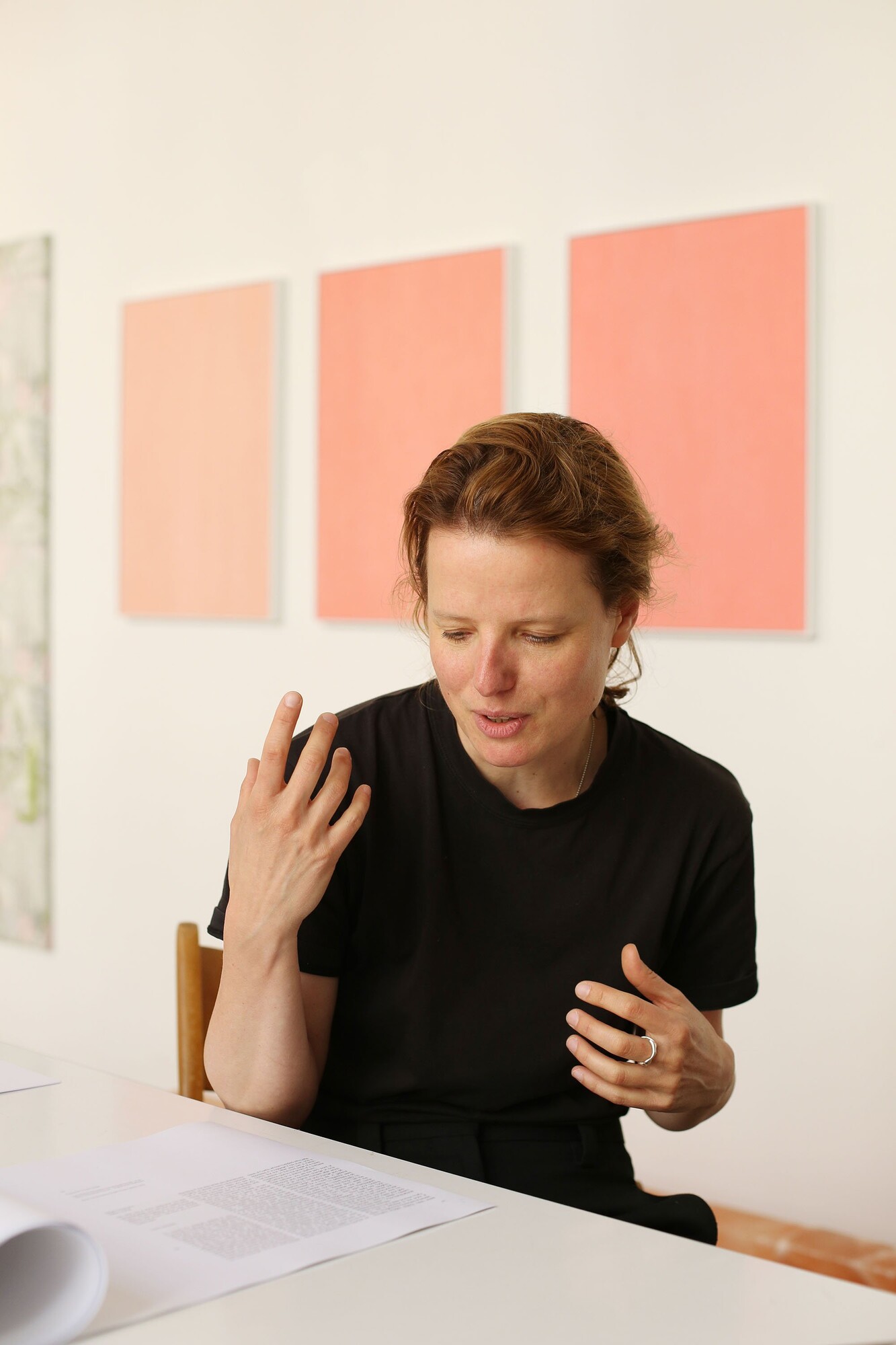 Person seated at a table in a studio, speaking with expressive gestures, with three large pink artworks on the wall behind them.